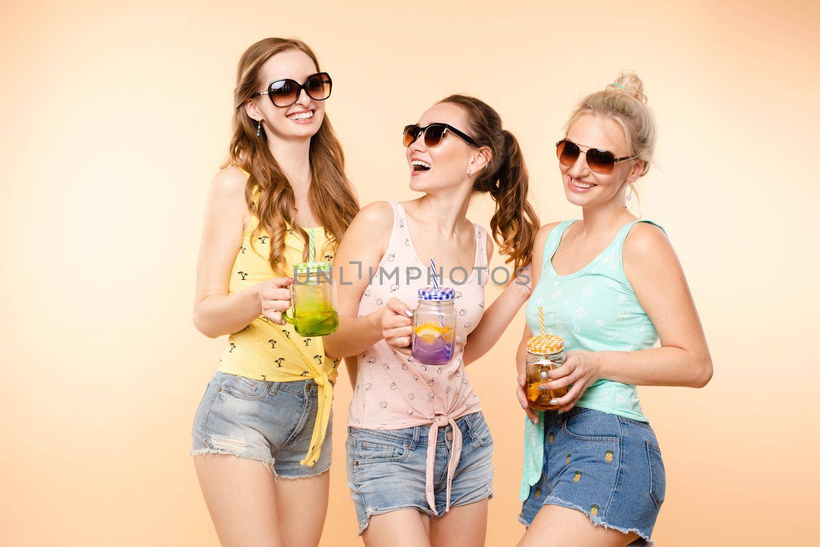 Three sporty girls in tops, shorts and glasses keeping fresh juice and posing on isolated background. Happy young women laughing and caring about body and fit. Concept of energy and shape.