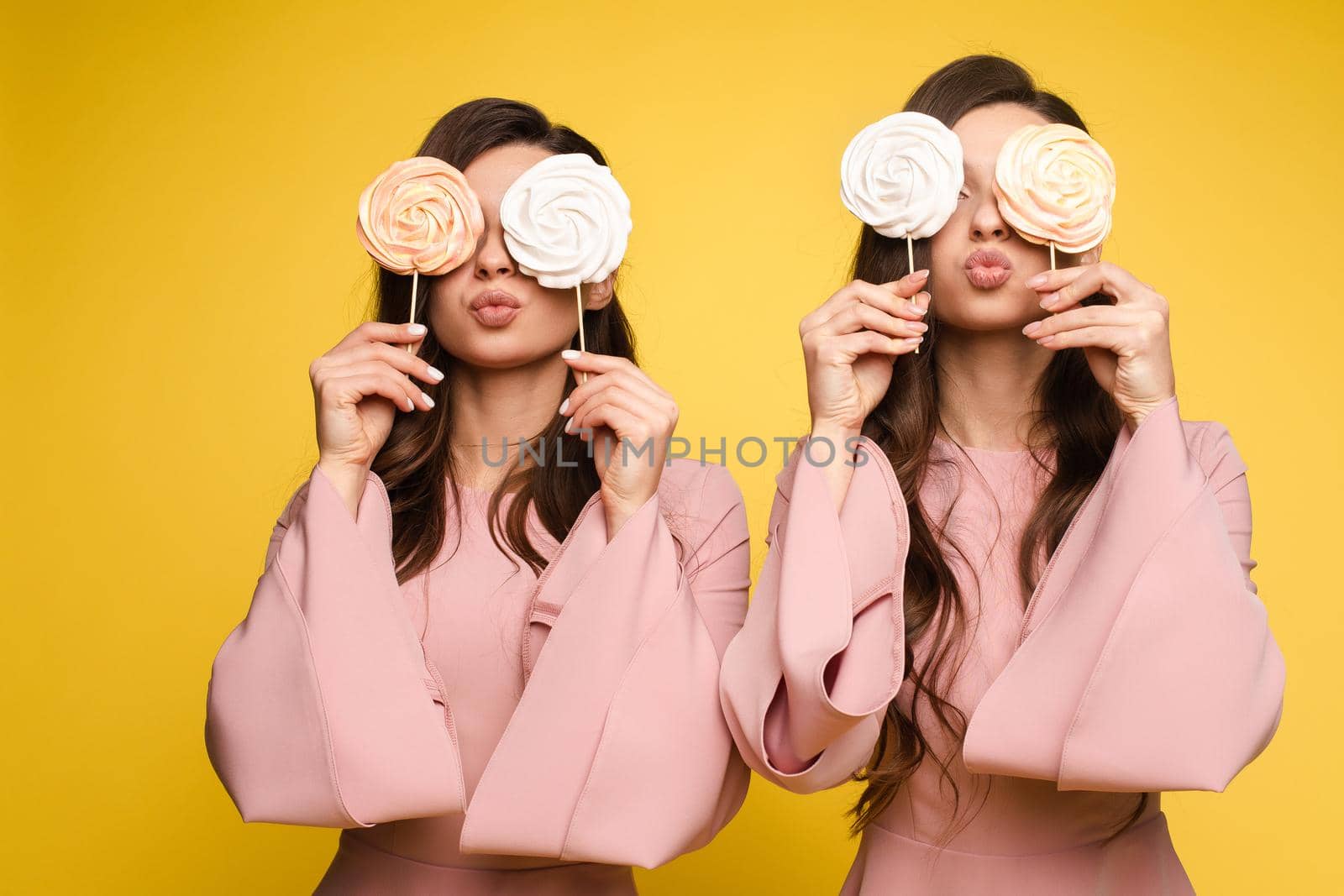 Front view of charming twins in pink dresses closing eyes with lollipops and posing on isolated background. Two female friends laughing and having fun in studio. Concept of childhood and beauty.