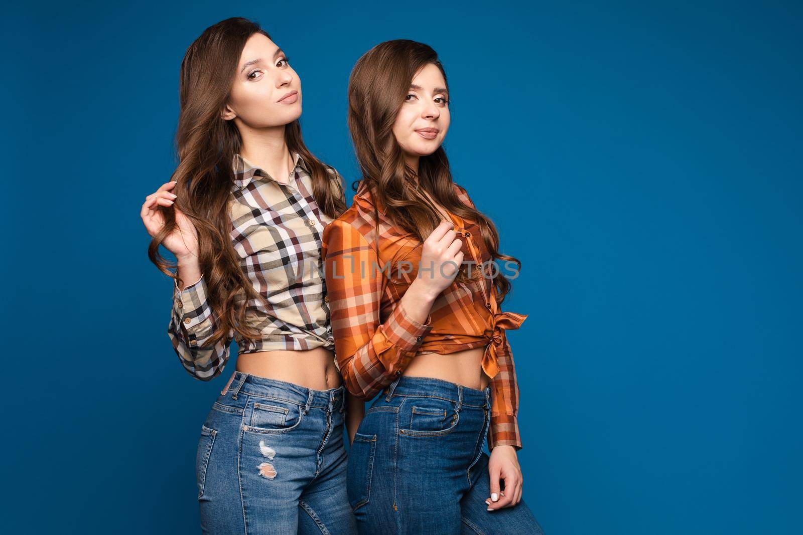 Two pretty slim sisters wearing checkered shirts, jeans and sneakers posing together on blue isolated background. Female sporty teens looking at camera and smiling. Concept of trends and style.