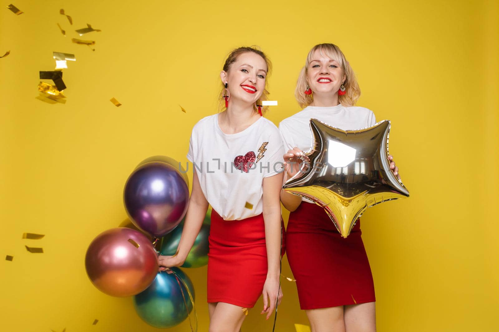 Two happy beautiful woman friend celebrating posing surrounded by colorful air balloon confetti by StudioLucky
