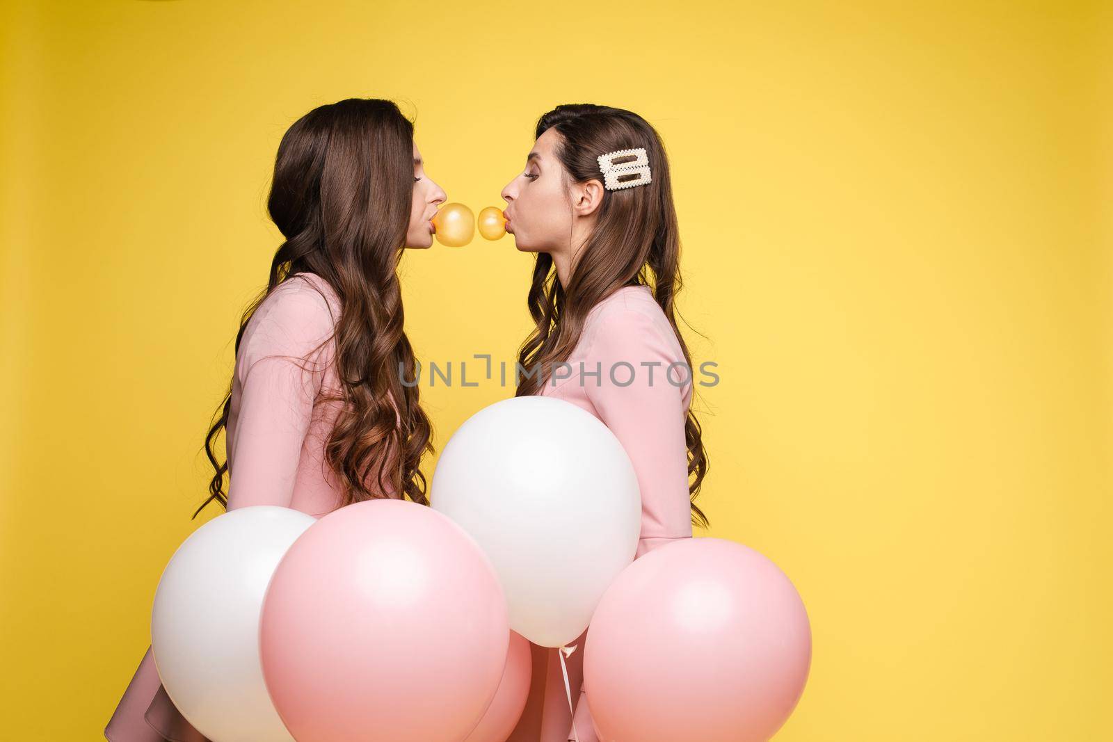 Young twins in elegant dresses holding pink and white balloons in their hands. Brunette sisters with long hair blowing big bubbles from chewing gum. Beautiful girls posing at camera with decorations.