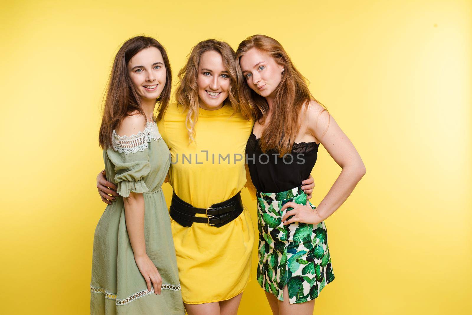 Three attractive young woman posing together isolated at yellow studio background medium long shot. Smiling fashionable young girl friend hugging enjoying friendship looking at camera