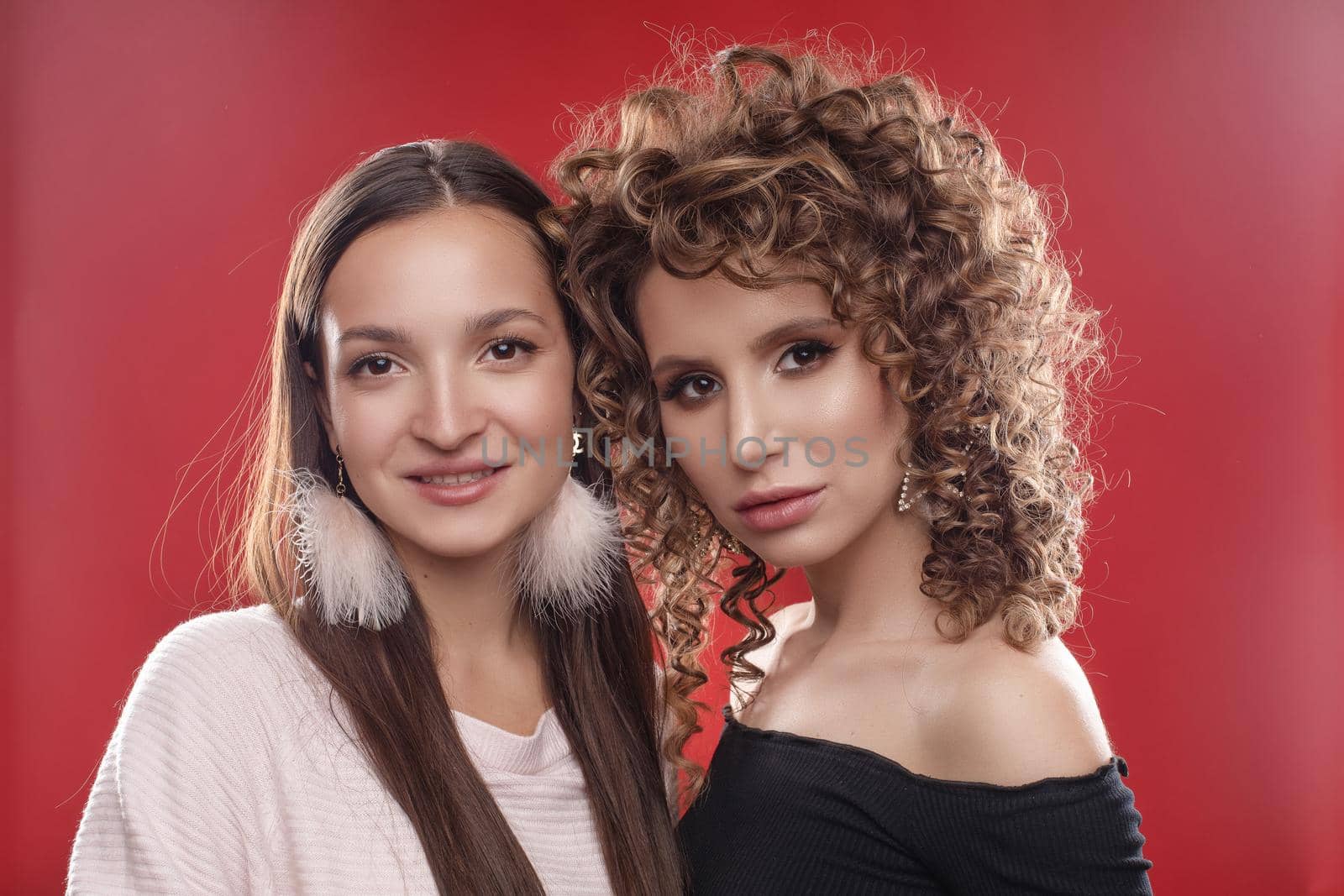Portrait of two beautiful glamour girls in black and white standing together. Young model with curly hair leaning on her friend's shoulder. Happy pretty lady with pink feather earrings smiling shyly.