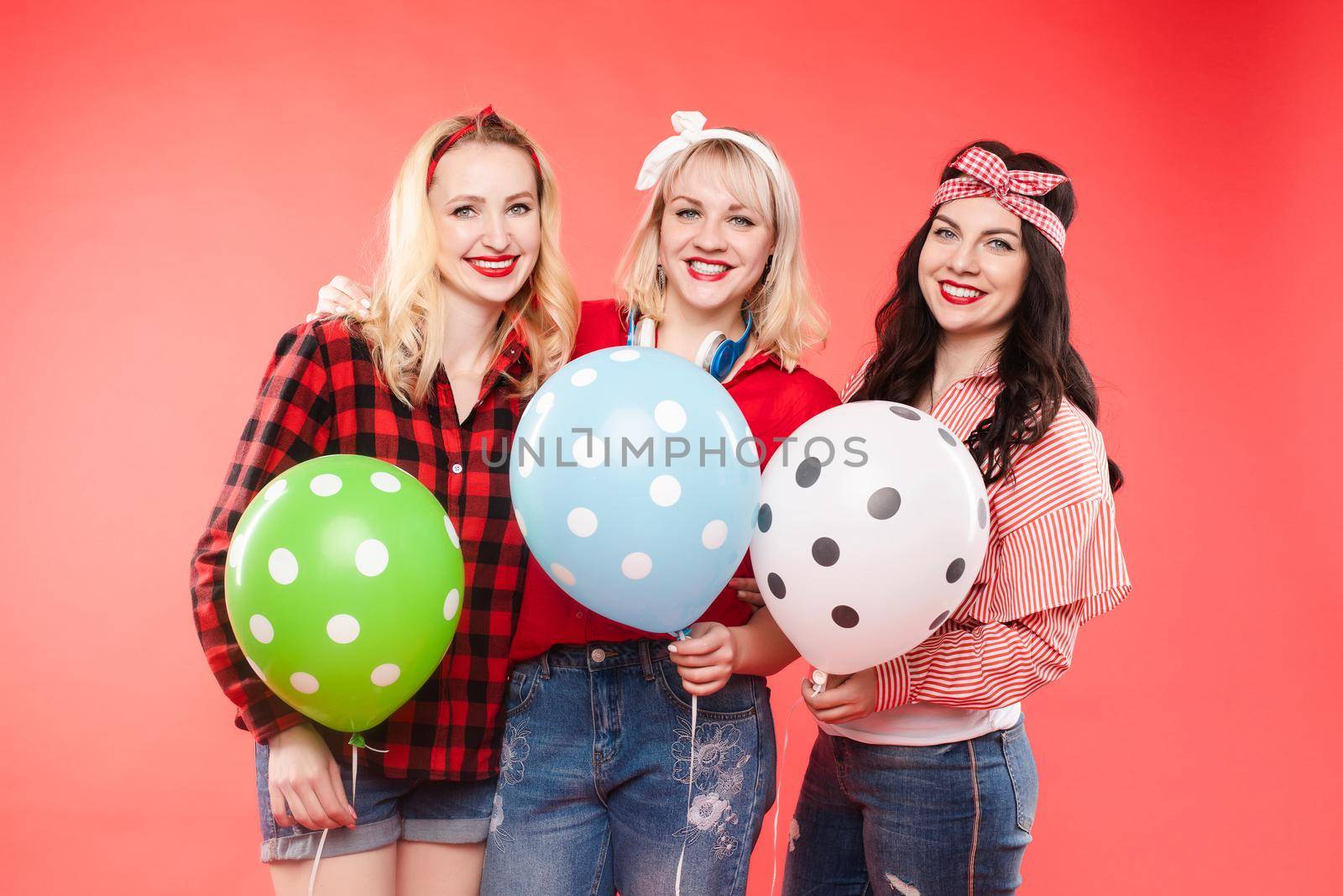 Stock photo of three cheerful beautiful and stylish girls hugging and smiling at camera. They are holding colorful dotted air balloons. Isolate on red background.