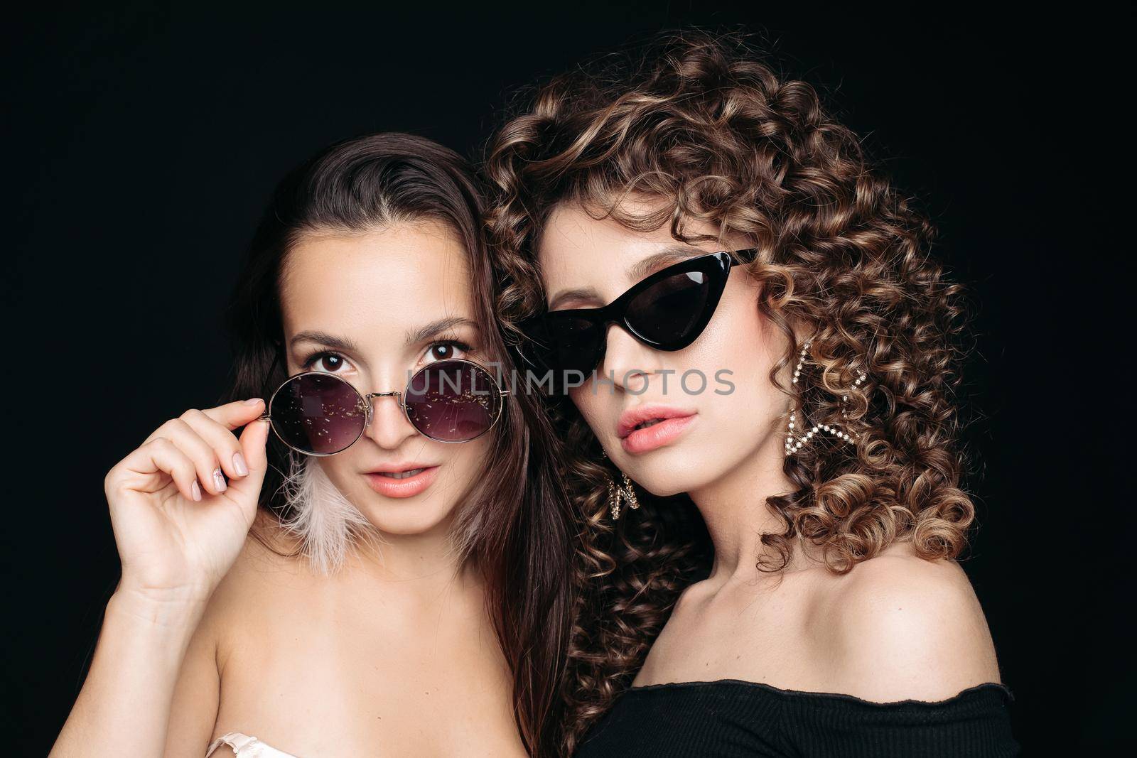 Front view of two different girls looking at camera and posing on black isolated background. Sexy brunette chewing gum while female friend laughing and keeping lollipop in hand. Concept of party.