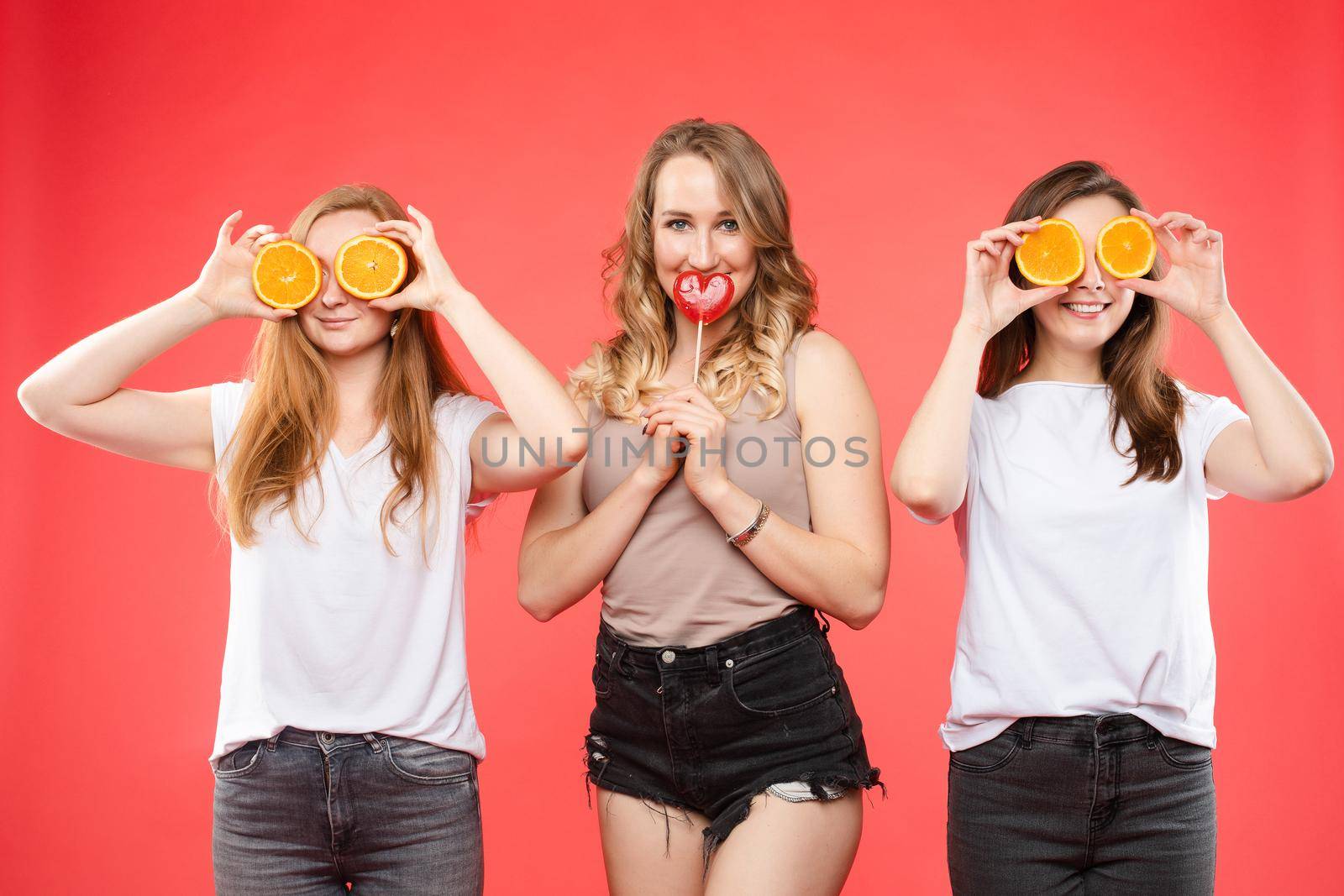 Stock photo of a beautiful girl with a red heart-shaped lollipop between two smiling girls with halved cut oranges. They are isolate on red background.