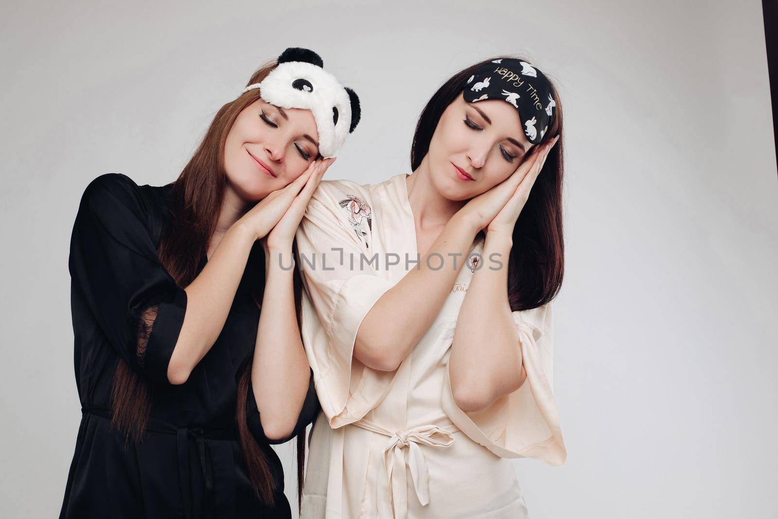 Two young tired girls wearing pajamas sleeping. Smiling models with closed eyes standing and keeping hands under heads and dreaming on white isolated background. Concept of bedtime and night.