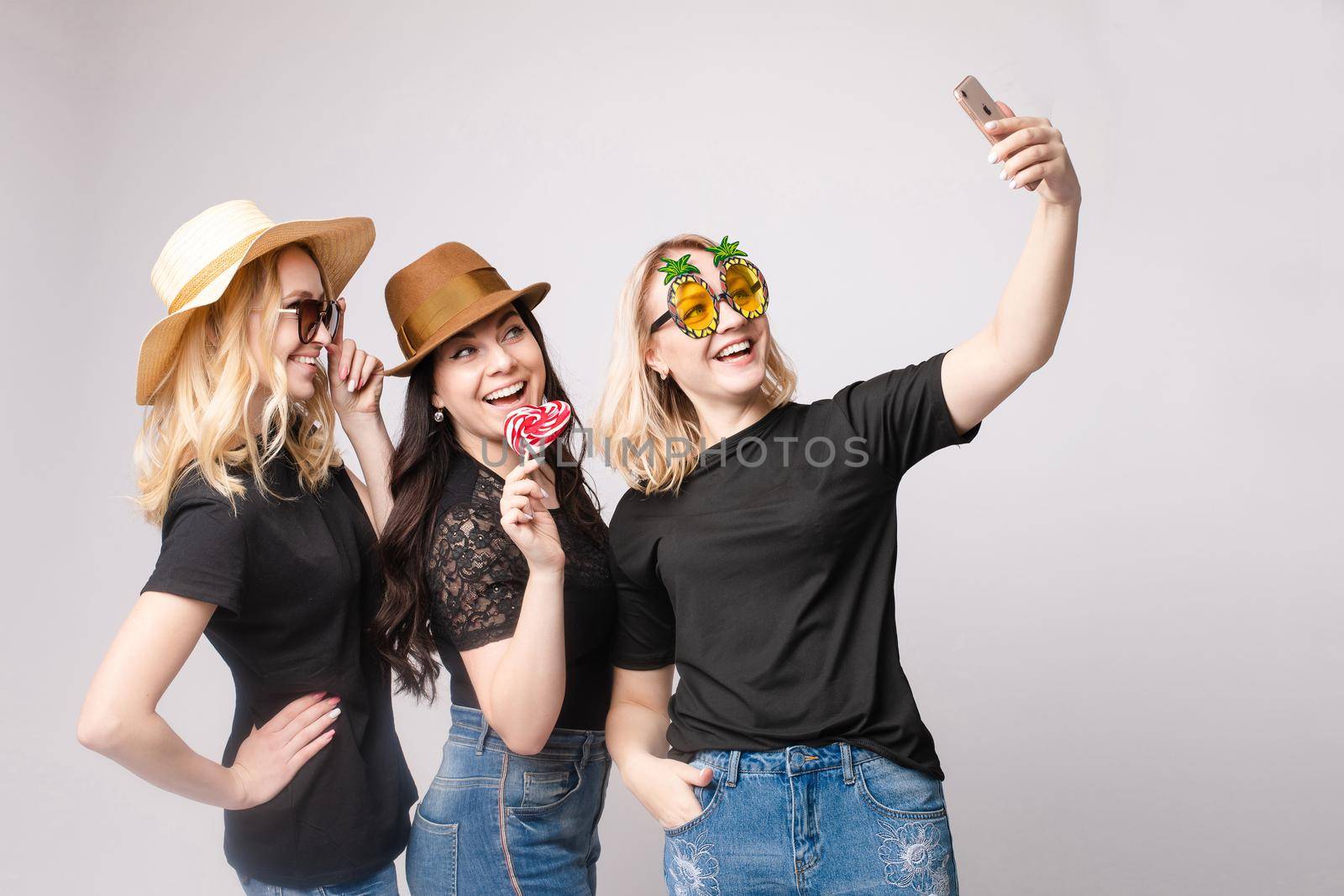 Smiling female friend wearing funny mask hat and eyeglasses posing taking selfie using smartphone medium shot. Happy woman having fun enjoying holiday party isolated at white studio background