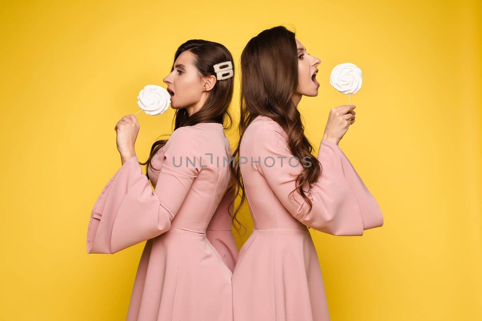 Front view of charming twins in pink dresses closing eyes with lollipops and posing on isolated background. Two female friends laughing and having fun in studio. Concept of childhood and beauty.