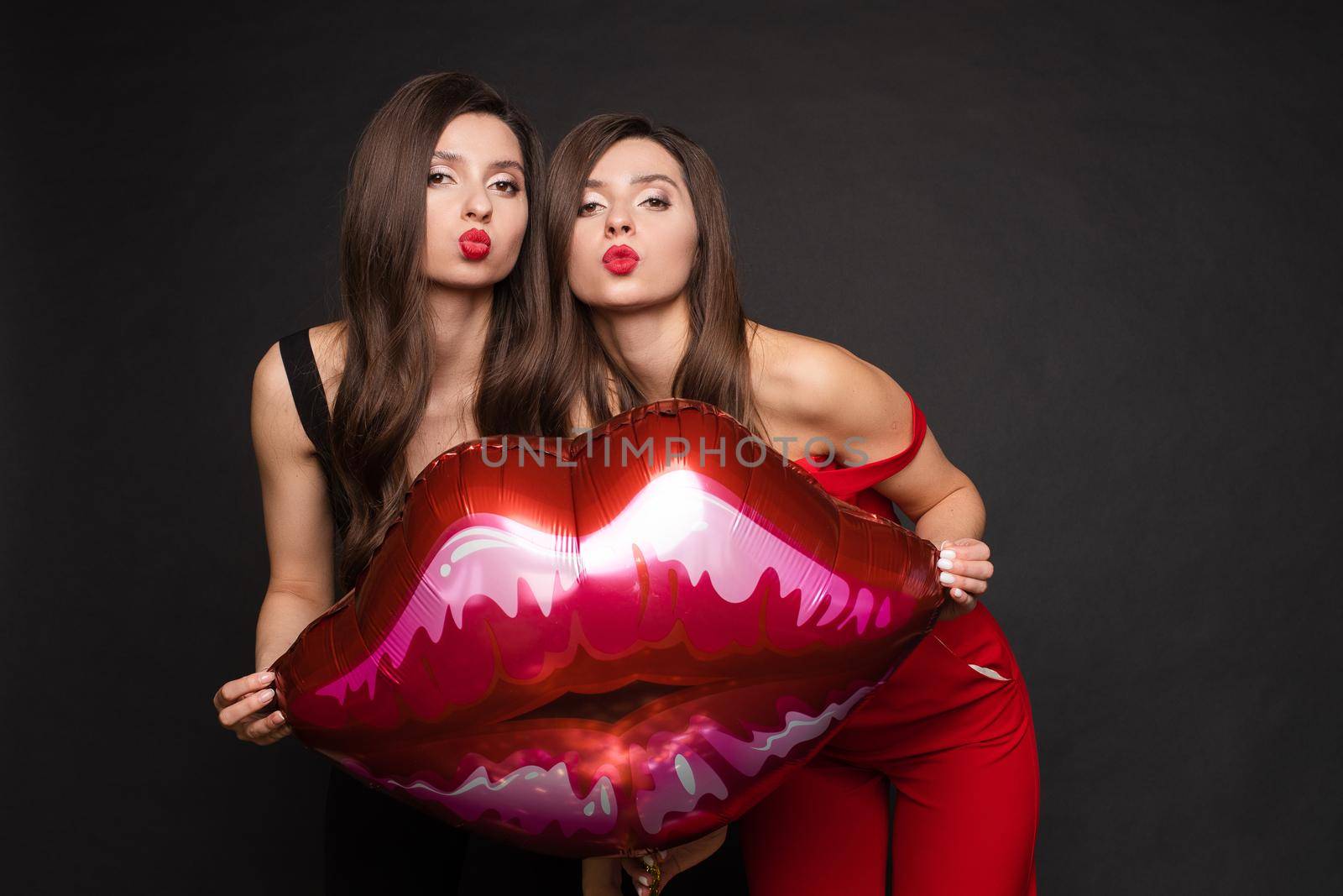 Close up of brunette twins with long hair in black and red costumes posing at camera. Young sisters standing together and holding red lips balloon. Gorgeous models with red lips sending air kisses.