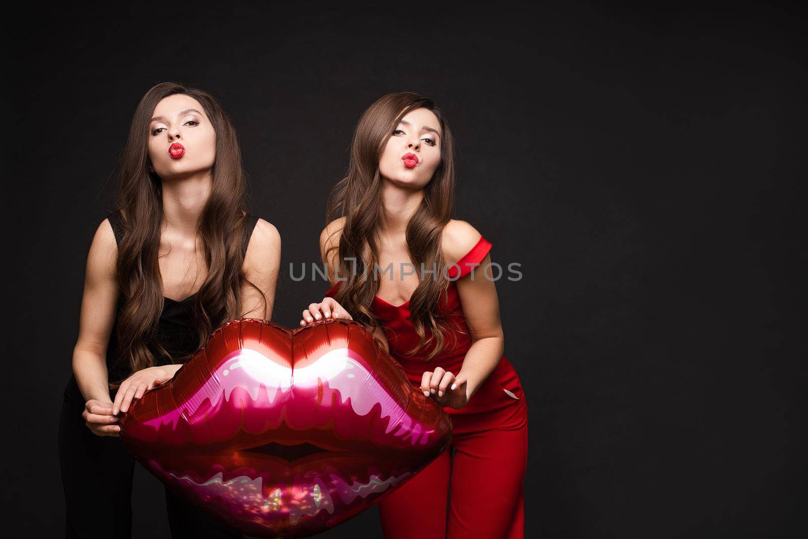 Close up of brunette twins with long hair in black and red costumes posing at camera. Young sisters standing together and holding red lips balloon. Gorgeous models with red lips sending air kisses.