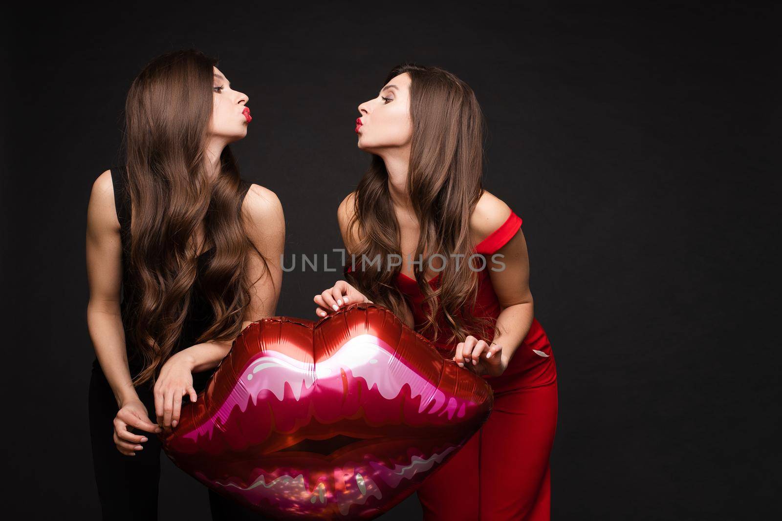 Close up of brunette twins with long hair in black and red costumes posing at camera. Young sisters standing together and holding red lips balloon. Gorgeous models with red lips sending air kisses.