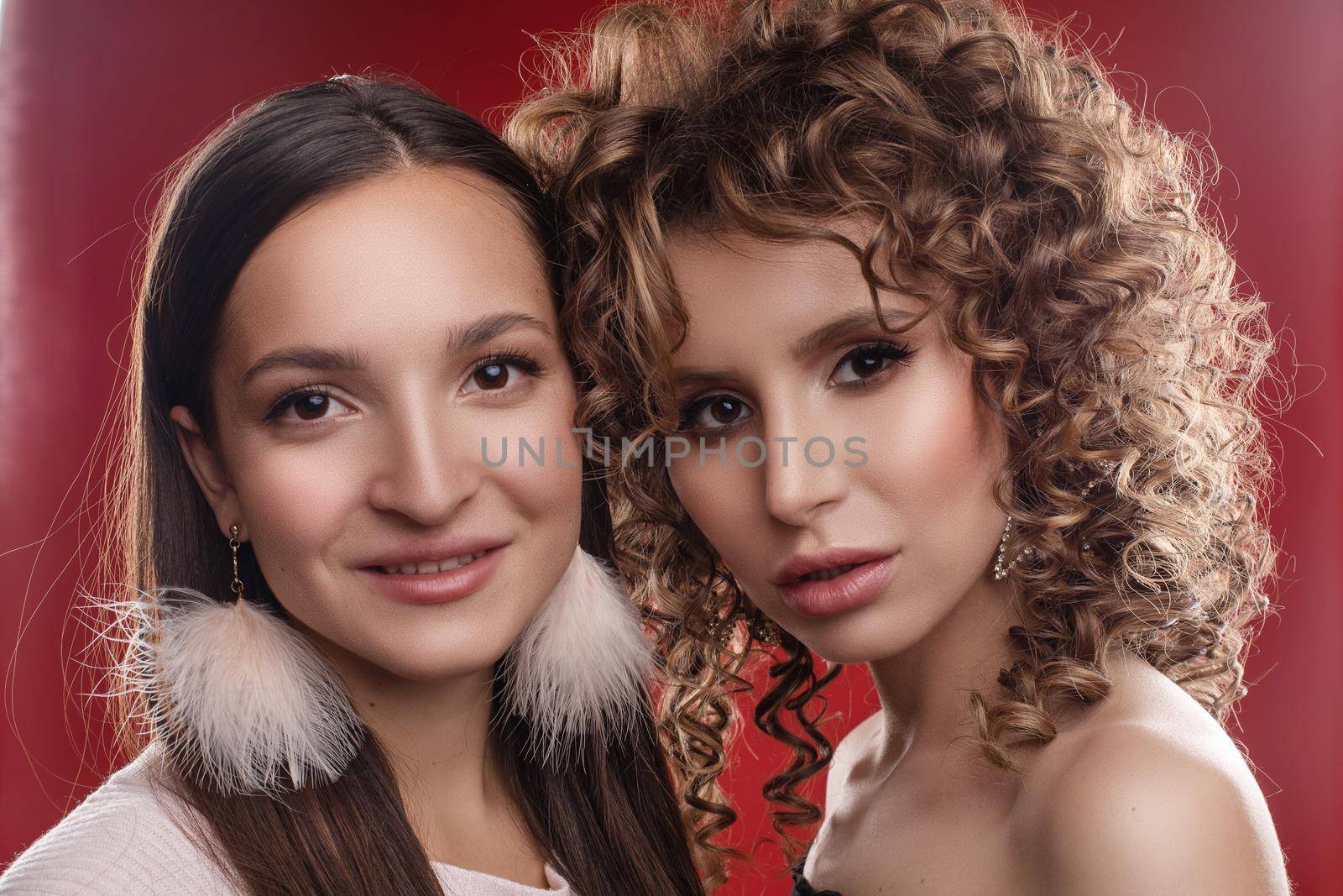 Front view of two different girls looking at camera and posing on red isolated background. Sexy brunette female friend. Concept of party.