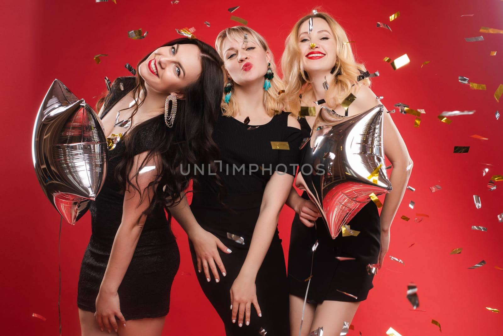 Group smiling woman posing surrounded by colorful shimmering confetti isolated red studio background by StudioLucky