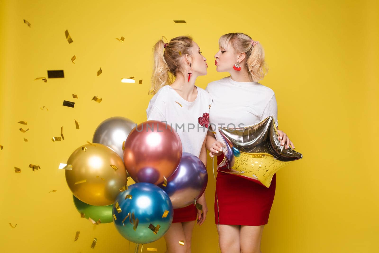 Two happy beautiful woman friend celebrating posing surrounded by colorful air balloon confetti. Attractive smiling female wearing bright clothes enjoying party isolated at yellow studio background