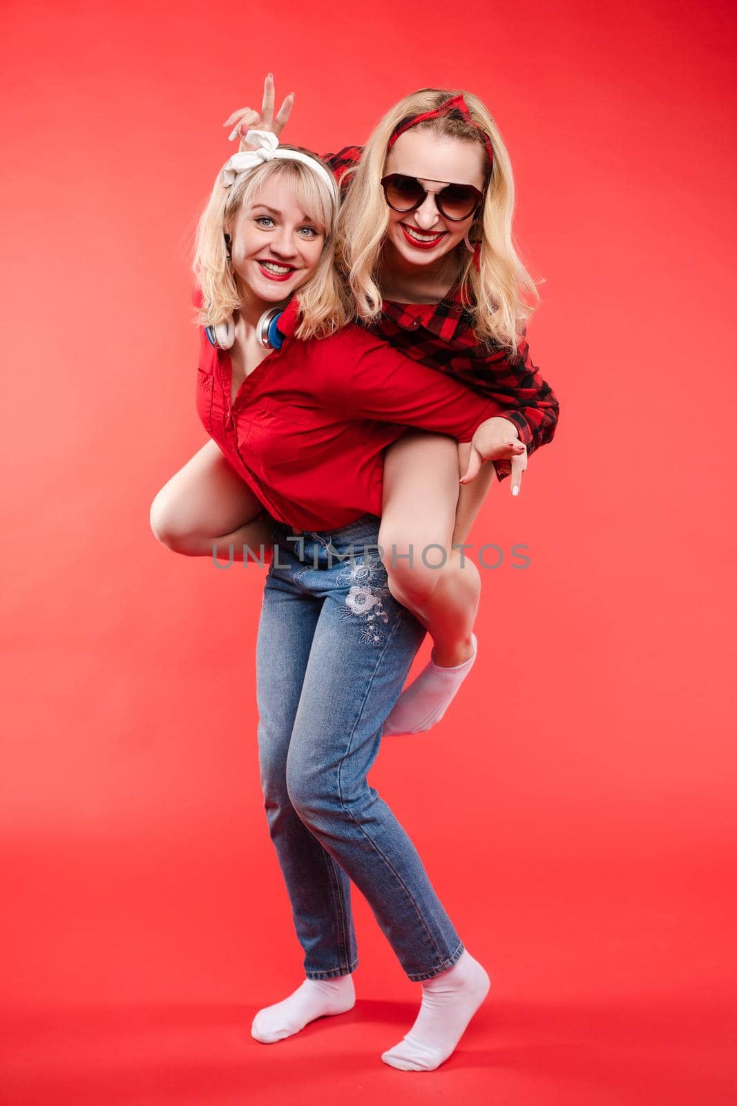 Stock studio portrait of two cheerful and positive trendy girlfriends or sisters having fun. Girl in sunglasses making bunny ears to her friend with her hand while sitting on her back. Isolate on red.