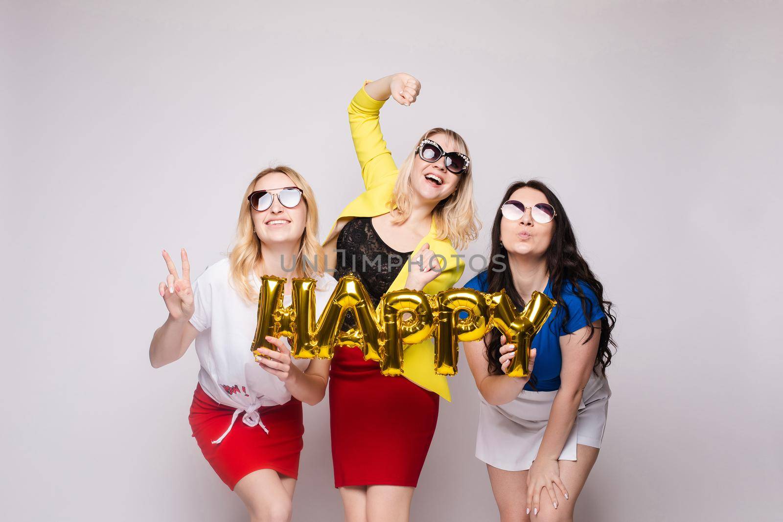Three happy girls keeping balloons, laughing and looking at camera on grey isolated background in studio. Cheerful women celebrating birthday party and posing. Concept of holiday and happiness.