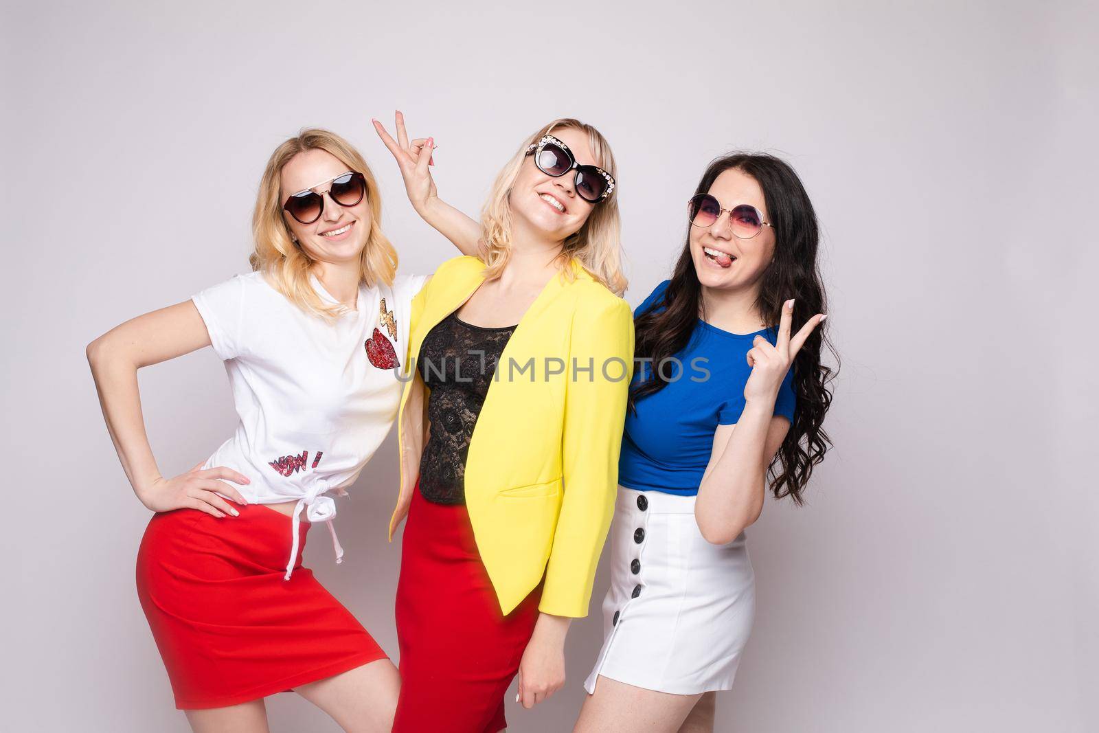 Three cheerful women wearing bright colorful skirts, shirts and glasses standing on grey isolated background. Happy girls looking at camera, laughing and showing tongue. Concept of happiness.