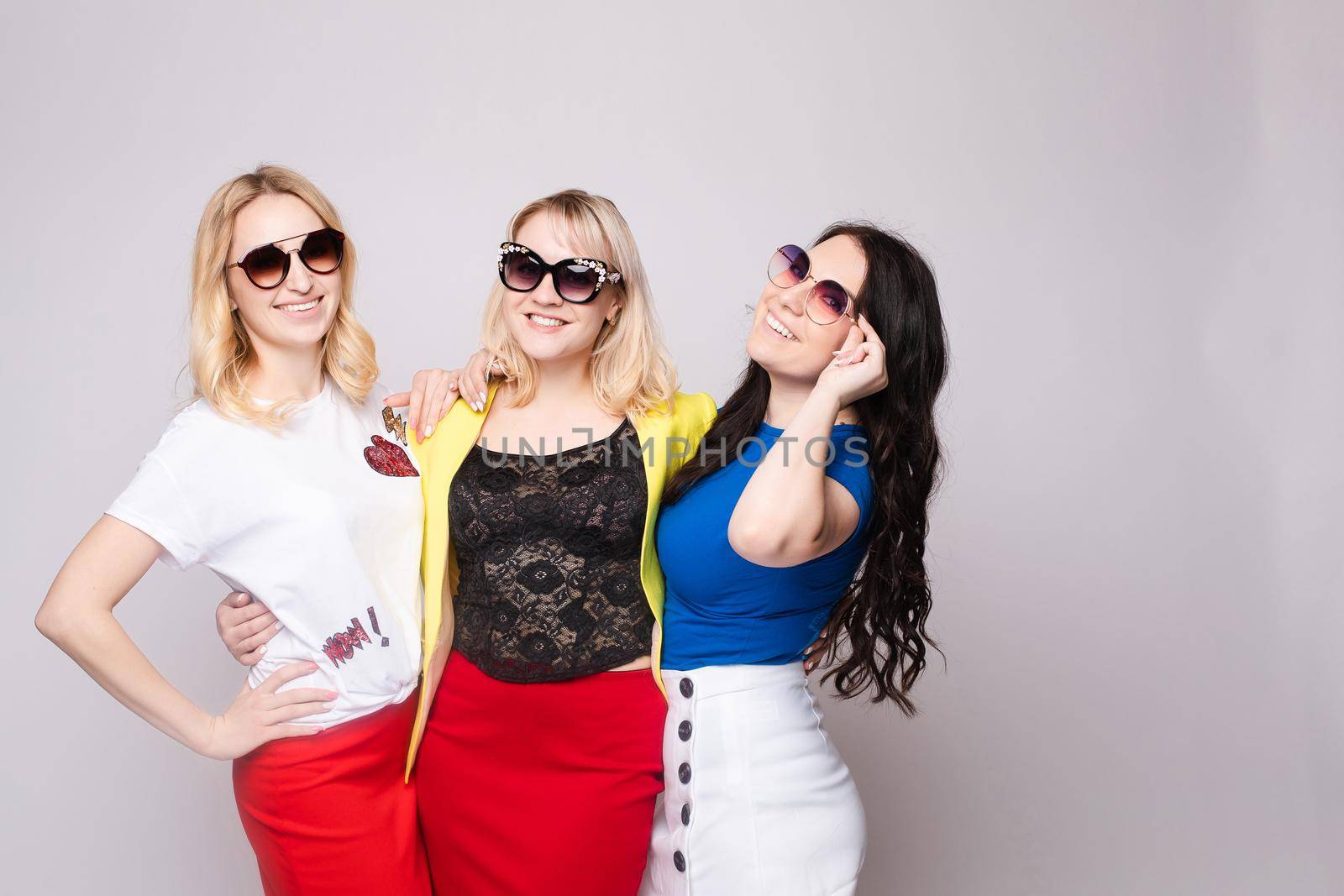 Three cheerful women wearing bright colorful skirts, shirts and glasses standing on grey isolated background. Happy girls looking at camera, laughing and showing tongue. Concept of happiness.
