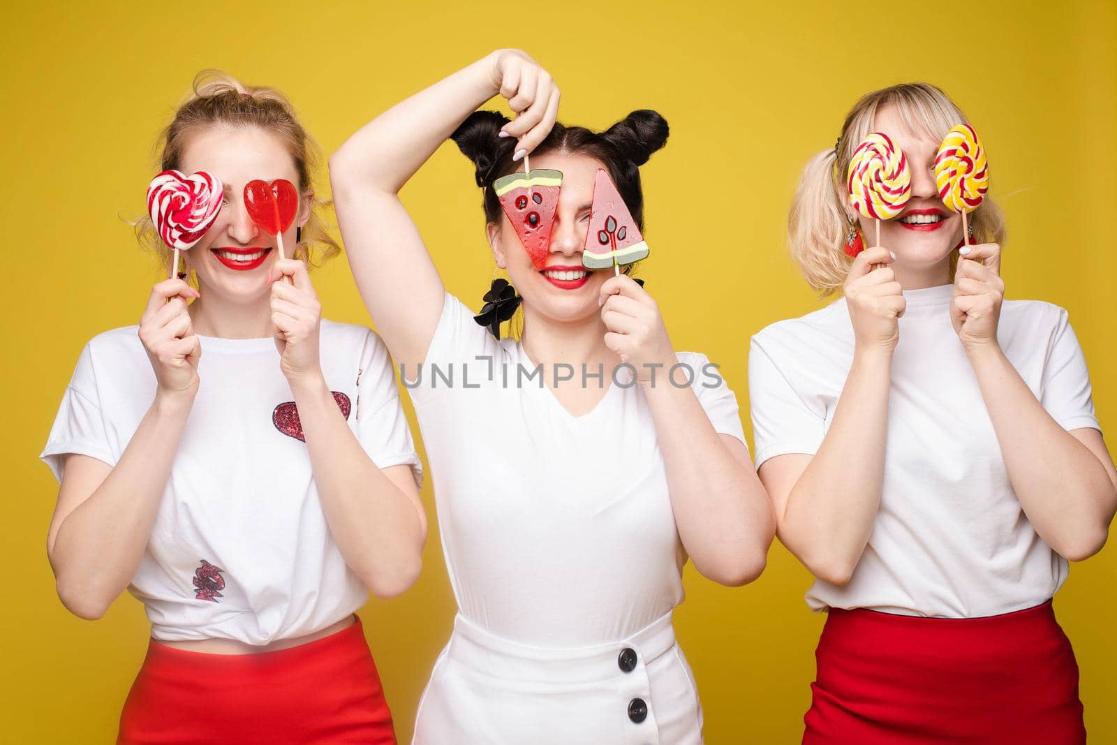 Group beautiful girl friend relaxing together posing with sweet colorful candy at yellow studio background. Happy smiling attractive woman having fun feeling rejoicing and happiness medium long shot