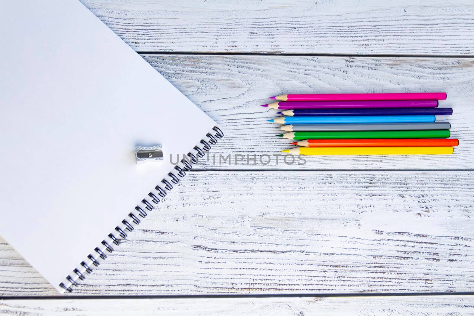 Bright, multicolored pencils for drawing in an album, a metal sharpener and a notebook with white sheets and a black spiral on a light, gray and wooden background.