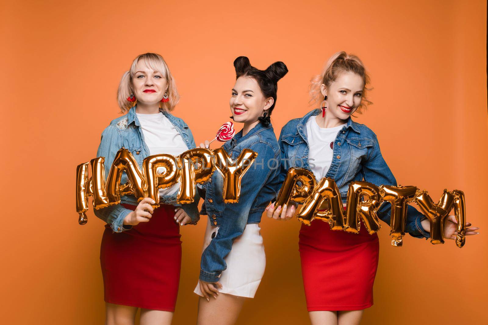 Front view of bright three girls in white shirts keeping balloons and ice creams on yellow isolated background. Cheerful pretty women looking at camera and posing. Concept of party and sparkles.