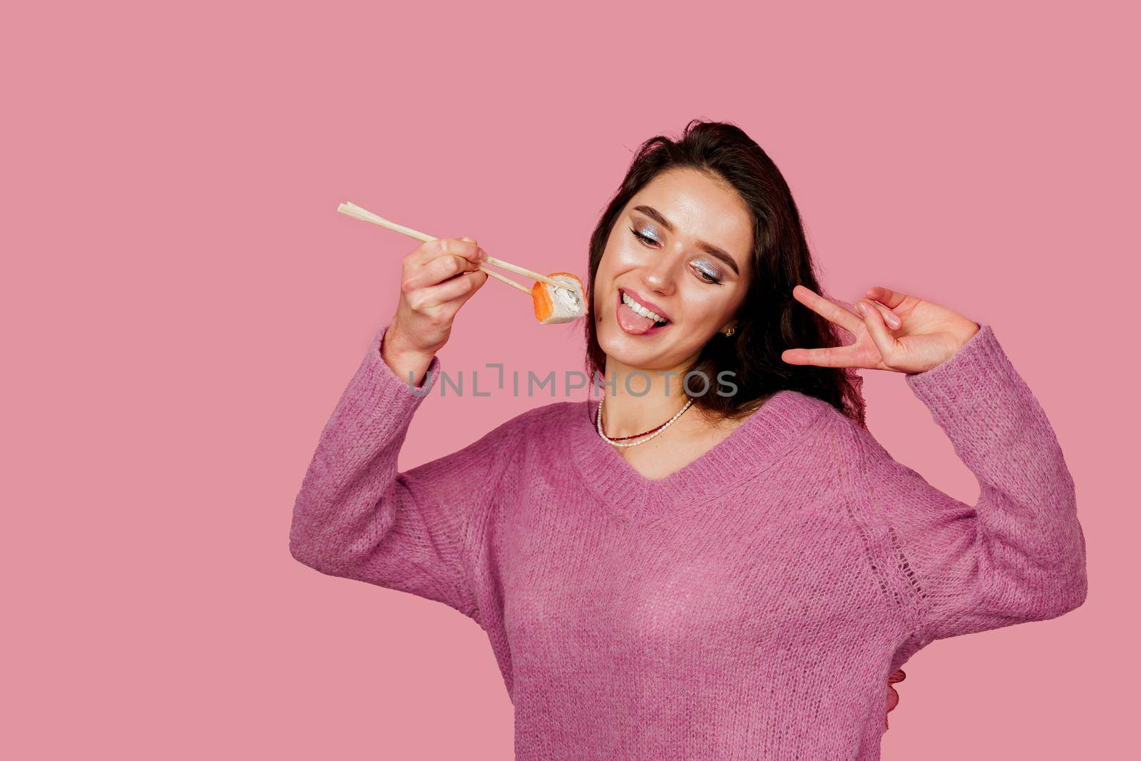 Happy girl with sushi on pink background. Young girl is holding sushi by chopsticks and wanting to eat one piece of roll philadelphia. Sushi delivery.