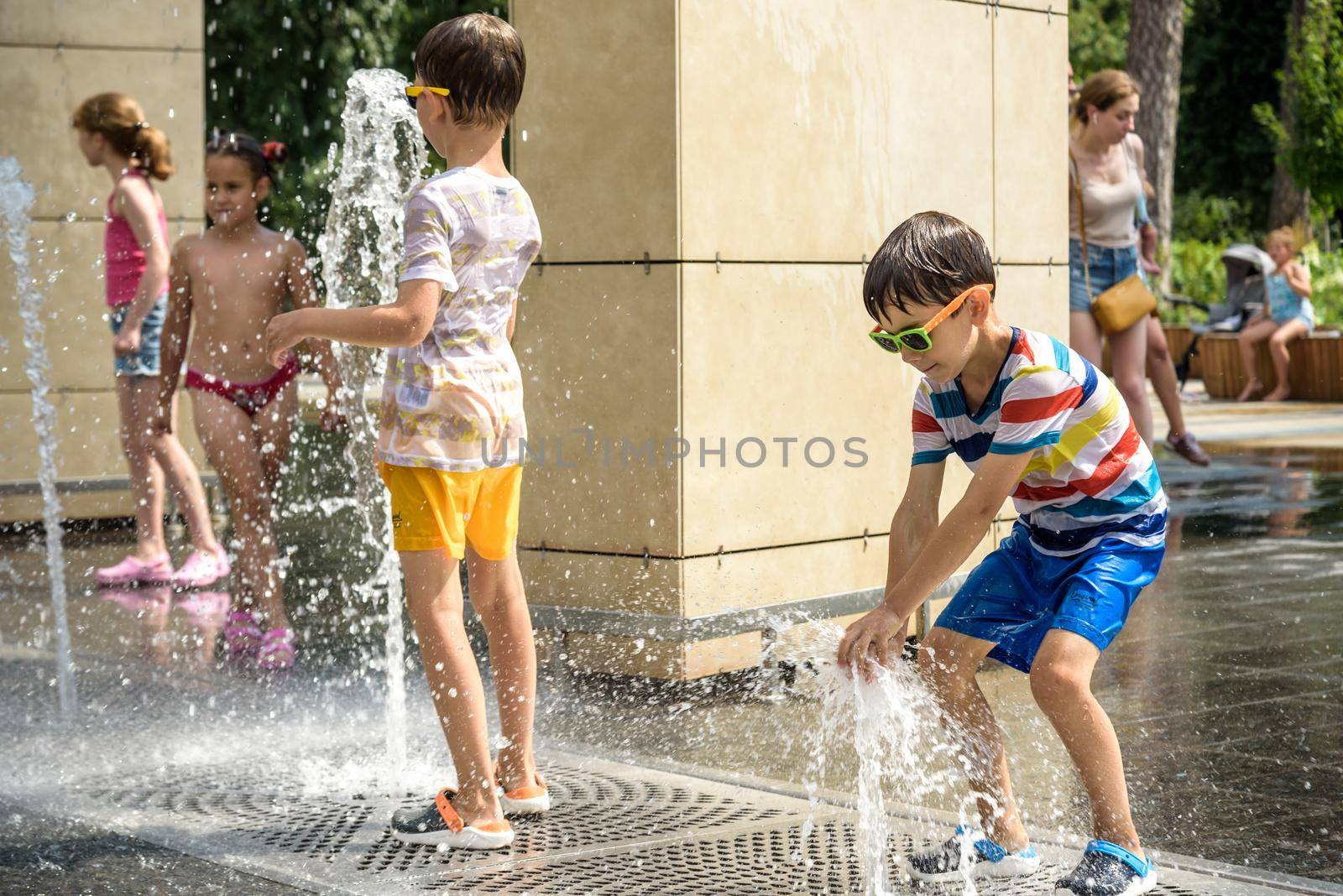 Kyiv, Ukraine - August 01, 2021: Boys jumping in water fountains. Children playing with a city fountain on hot summer day. Happy friends having fun in fountain. Summer weather. Friendship, lifestyle and vacation.