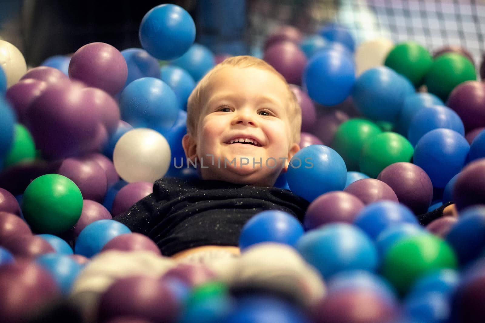 Blonde little boy lying on multi coloured plastic balls in big dry paddling pool in playing centre. Smiling at camera by Lincikas