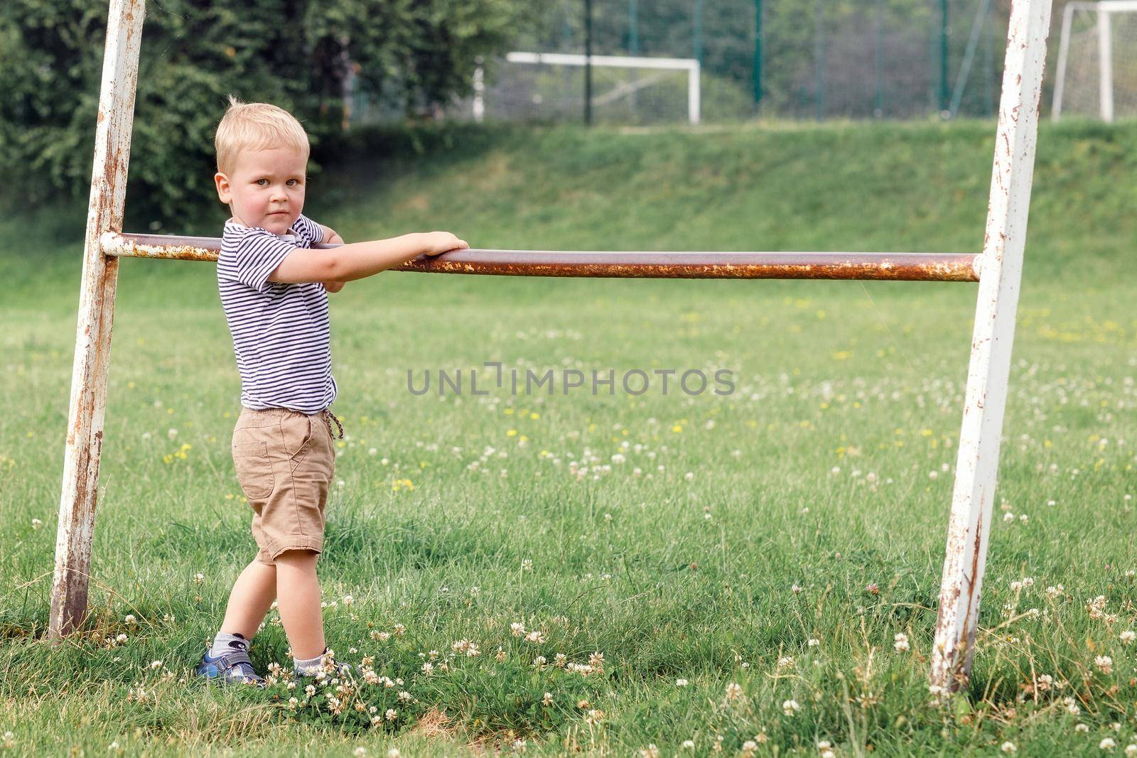 Cute kid posing and looking at camera. Holding old iron railing in a greenfield background.