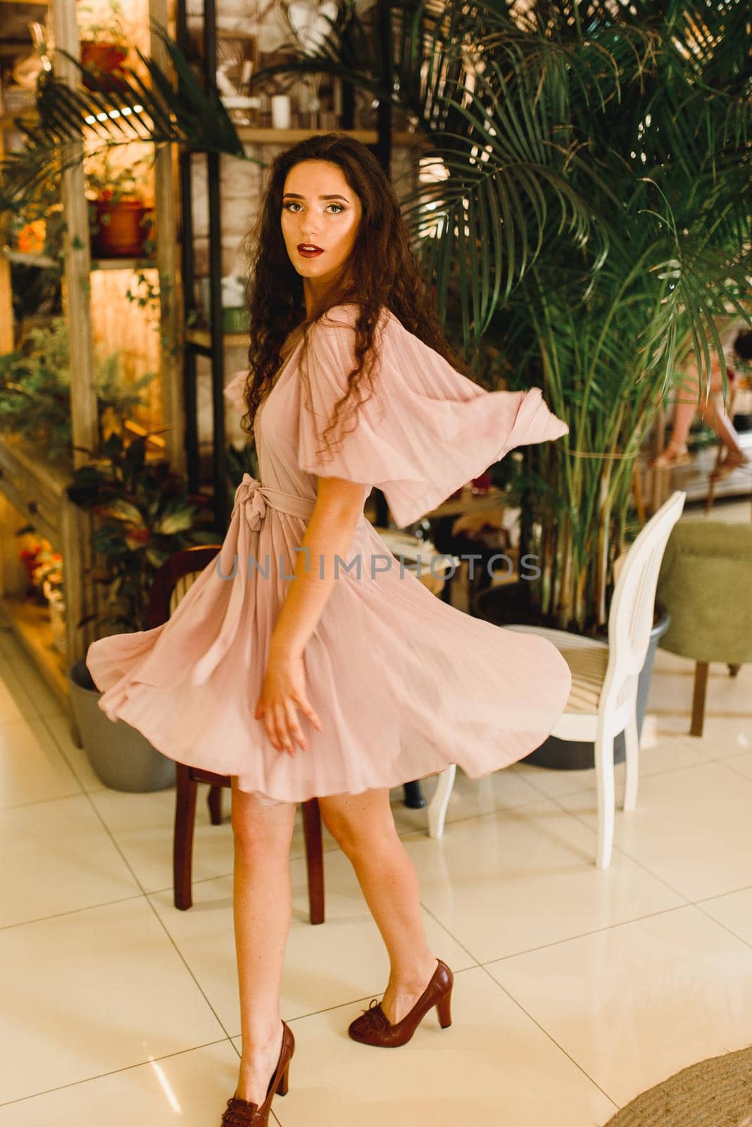 Young woman in soft pink dress is moving and looking into the camera. Beauty girl in cafe with big green plants