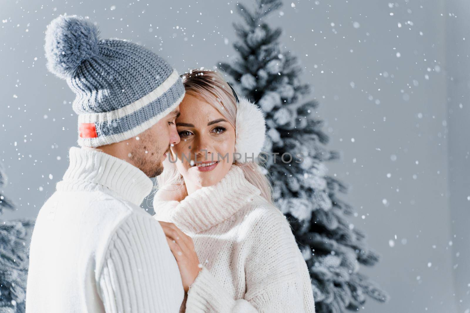 Happy young couple hugs and smiles and snow falls near christmas trees at the eve of new year celebration in winter day. Smiley man and woman weared white pullovers love each other