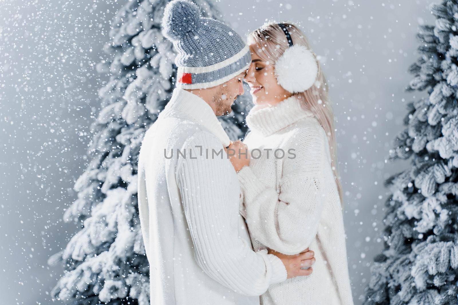 Happy young couple hugs and smiles and snow falls near christmas trees at the eve of new year celebration in winter day. Smiley man and woman weared white pullovers love each other, by Rabizo