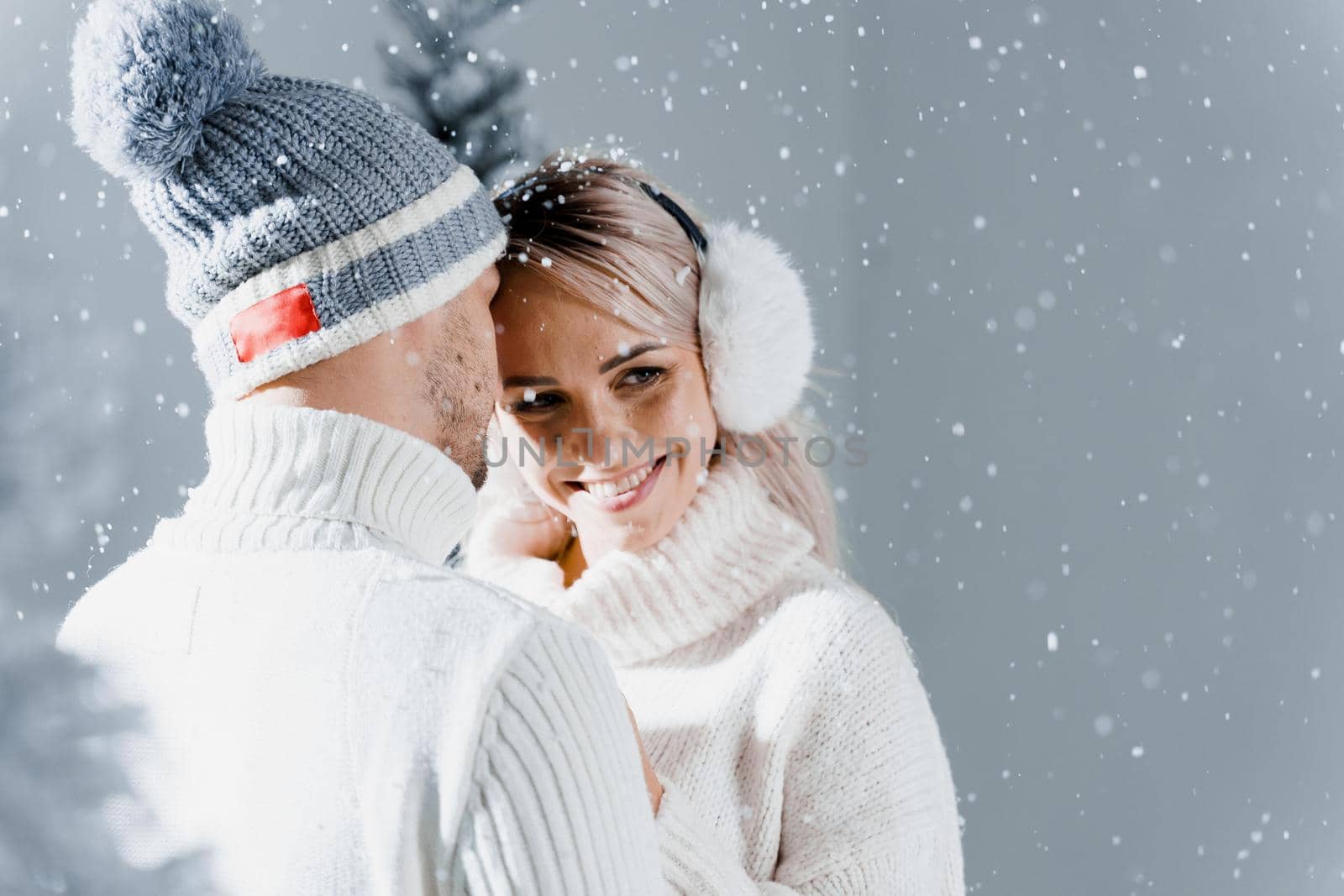 Happy young couple hugs and smiles and snow falls near christmas trees at the eve of new year celebration in winter day. Smiley man and woman weared white pullovers love each other, by Rabizo