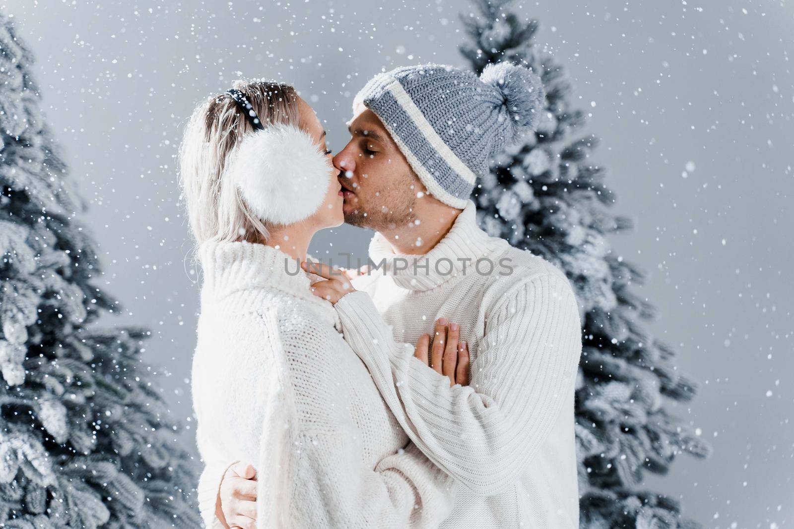 Winter love story with couple weared fur headphones, hats, white sweaters. Happy young couple hugs and kiss near christmas trees at the eve of new year celebration in winter day by Rabizo
