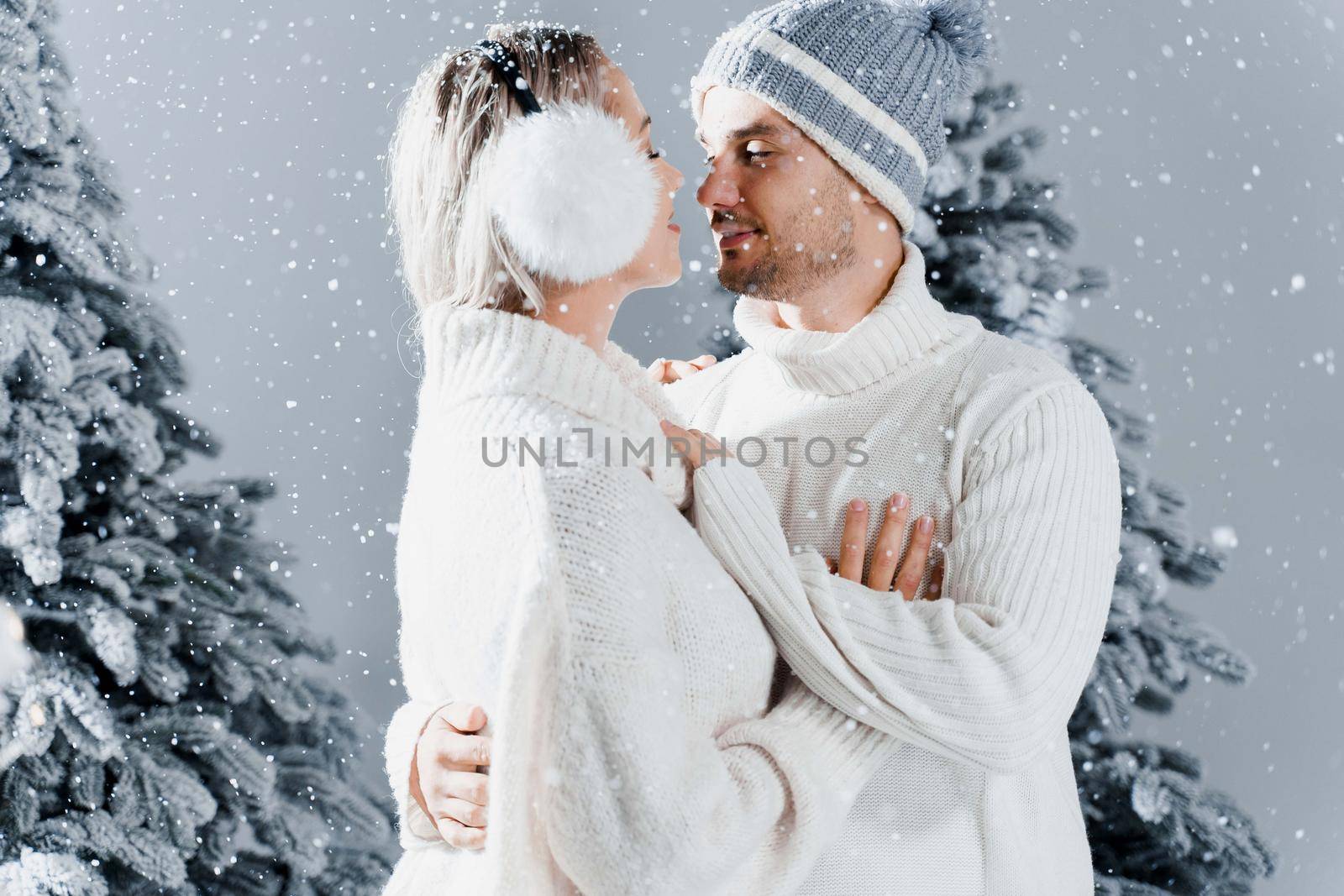 Winter love story with couple weared fur headphones, hats, white sweaters. Happy young couple hugs and kiss near christmas trees at the eve of new year celebration in winter day.