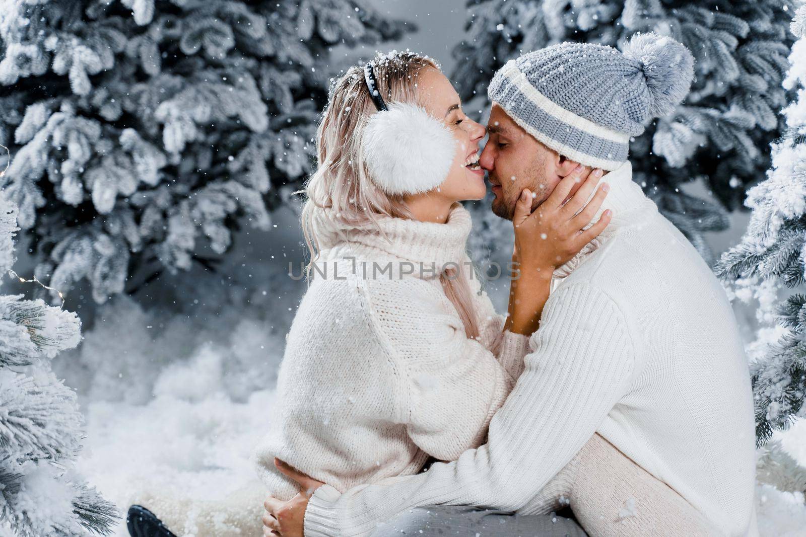 Man kiss and hug his woman and snow falls. New year love story. People weared wearing fur headphones, hats, white sweaters by Rabizo