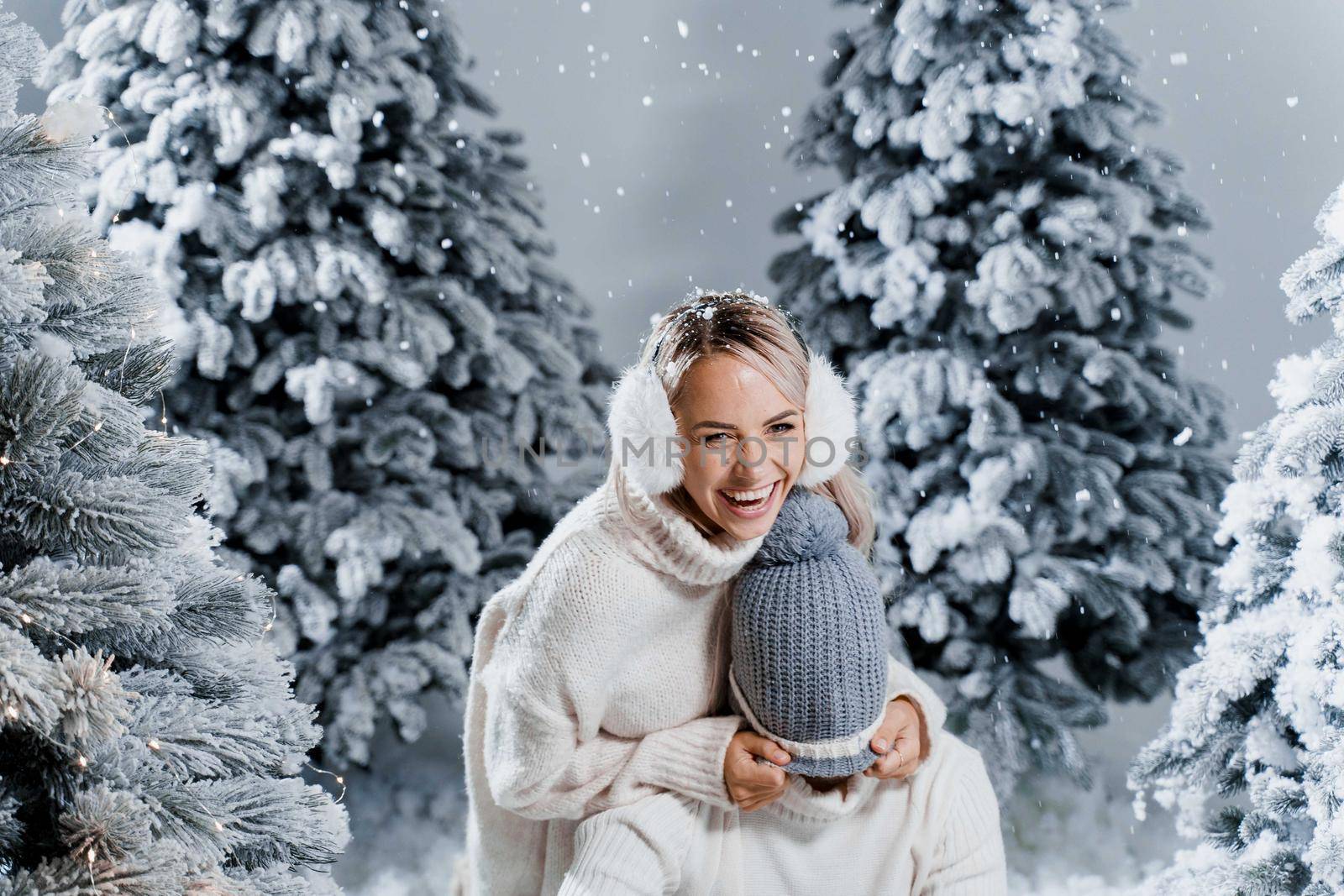 Falling snow and kisses. Happy young couple close-up hugs and kiss near christmas trees at the eve of new year celebration in winter day. Smiley man and woman weared white pullovers love each other, by Rabizo