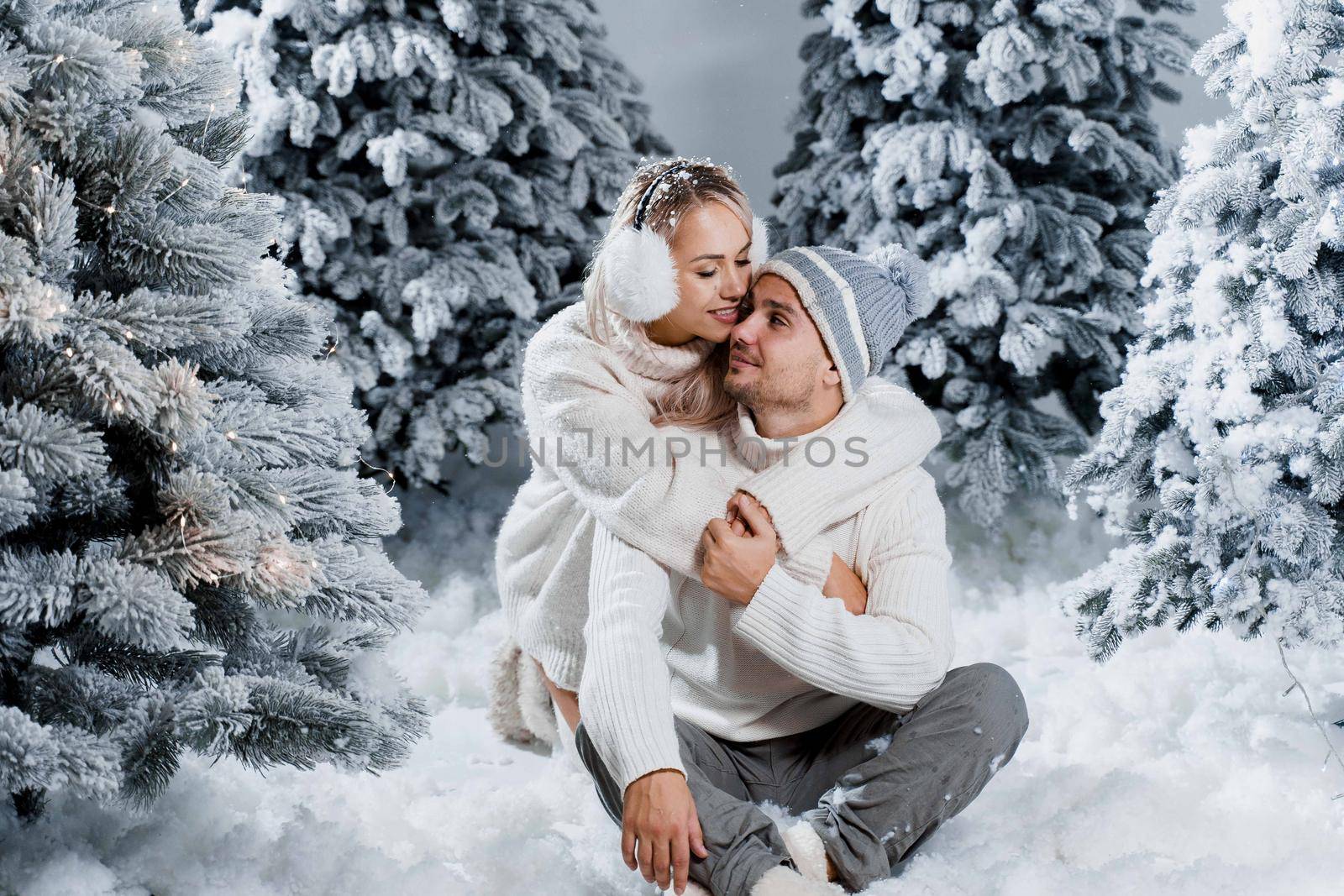 New year love story. Couple kisses and hugs. People weared wearing fur headphones, hats, white sweaters. Happy young couple hugs and kiss near christmas trees in winter day by Rabizo