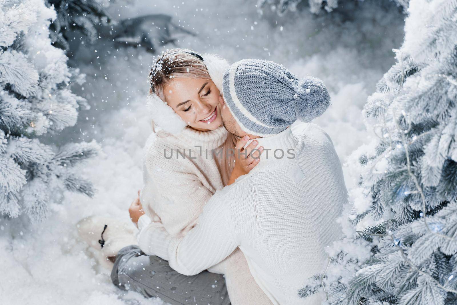 Man kiss and hug his woman and snow falls. New year love story. People weared wearing fur headphones, hats, white sweaters.
