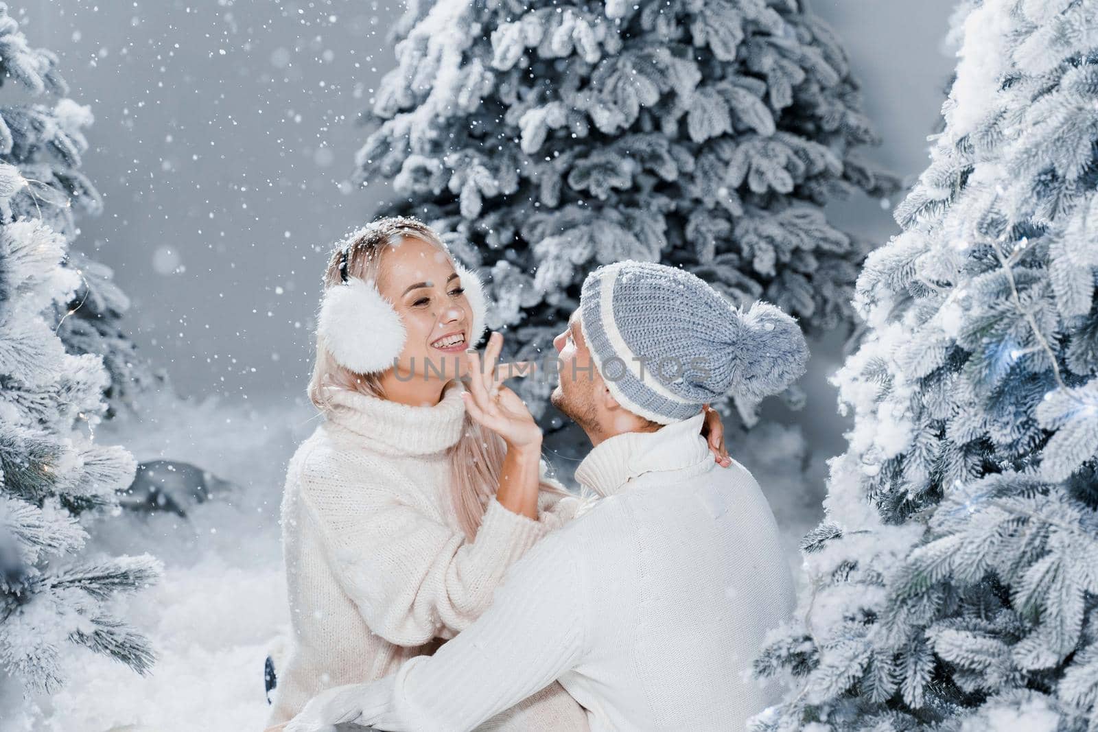 Couple seat on the snow and hug, kiss, and have fun each other. Winter love story before new year celebration. Waiting for christmas gift. Happy couple weared fur headphones, hats, white sweaters.