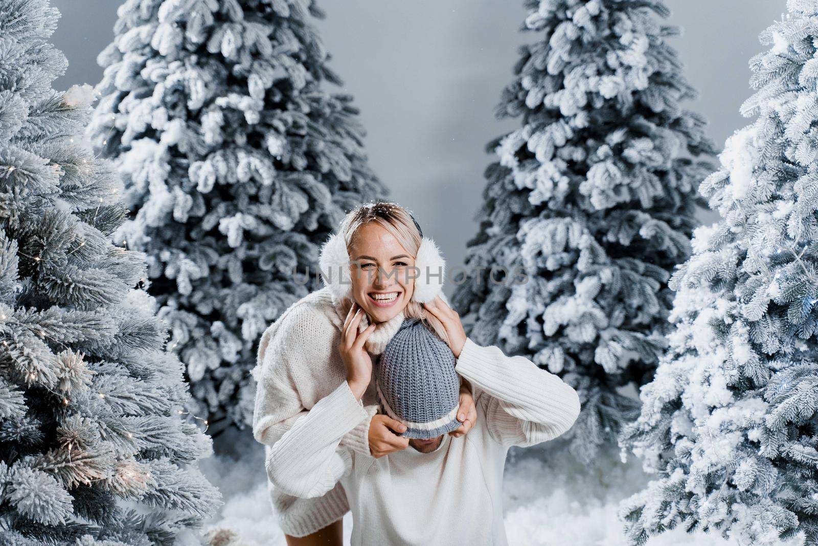 Falling snow and kisses. Happy young couple close-up hugs and kiss near christmas trees at the eve of new year celebration in winter day. Smiley man and woman weared white pullovers love each other