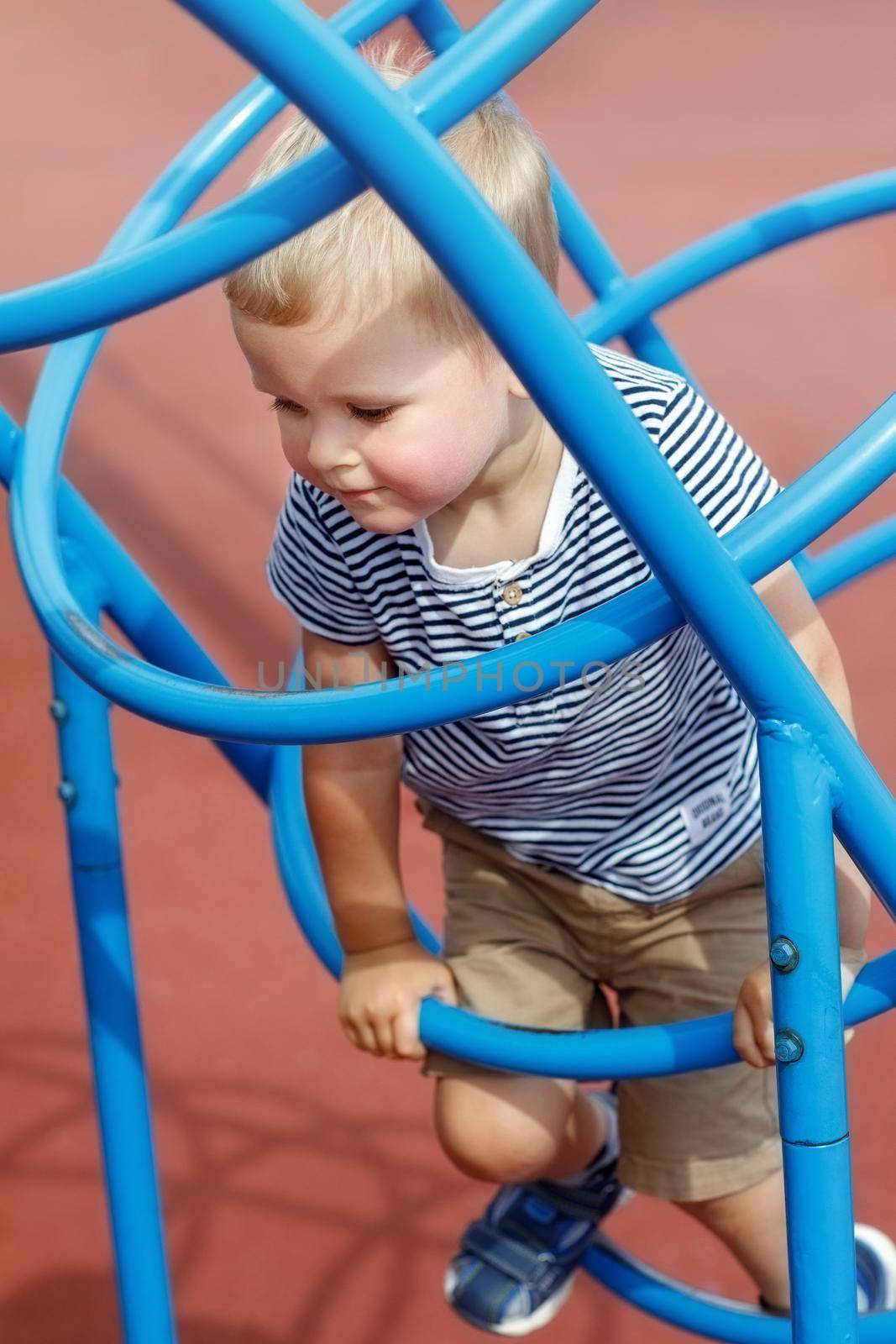 Focused boy climbs on the blue, tube shaped playground ladder. Frisky kid enjoying sunny day in a public. Does not  care  the risk