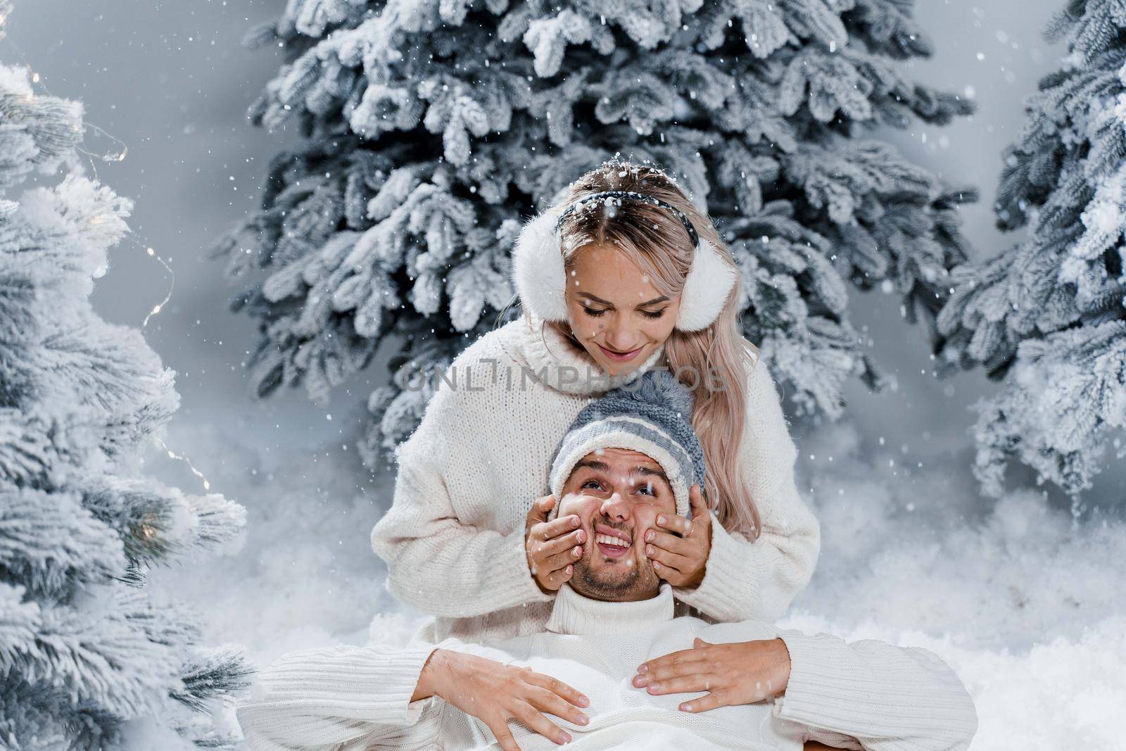 Falling snow and kisses. Happy young couple close-up hugs and kiss near christmas trees at the eve of new year celebration in winter day. Smiley man and woman weared white pullovers love each other