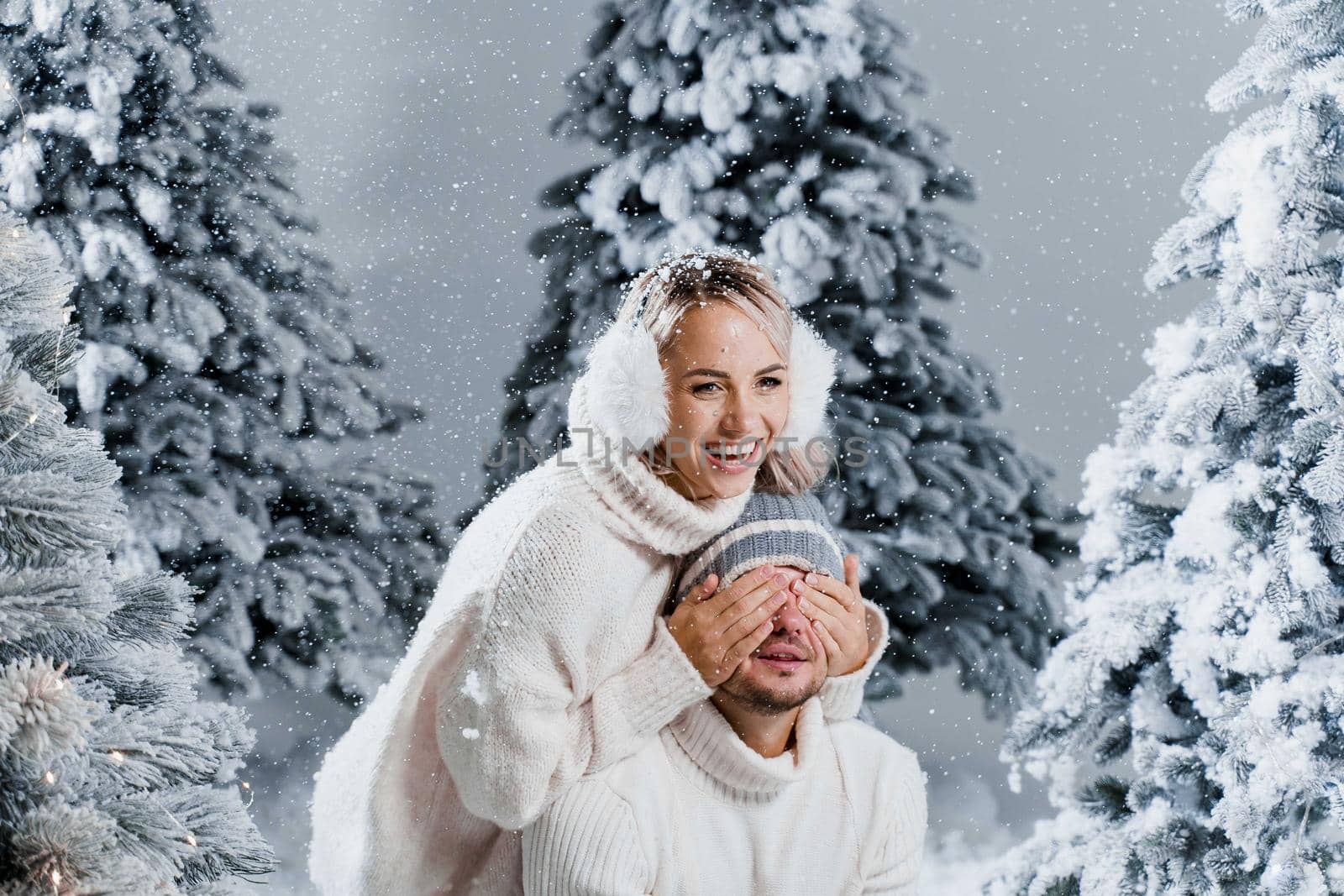 Winter love story with couple weared fur headphones, hats, white sweaters. Happy young couple hugs and kiss near christmas trees at the eve of new year celebration in winter day.