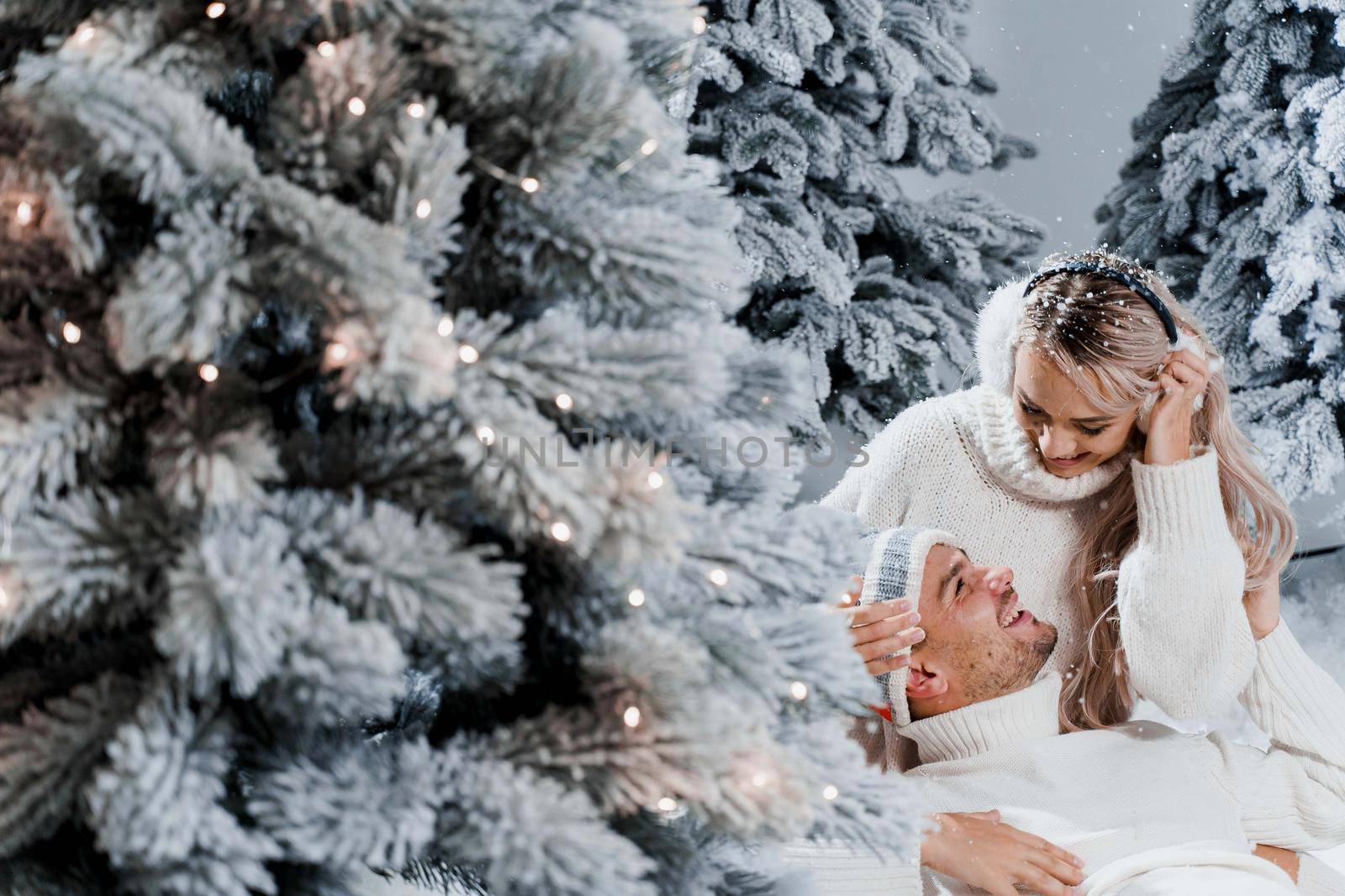 New year love story. Couple kisses and hugs. People weared wearing fur headphones, hats, white sweaters. Happy young couple hugs and kiss near christmas trees in winter day.