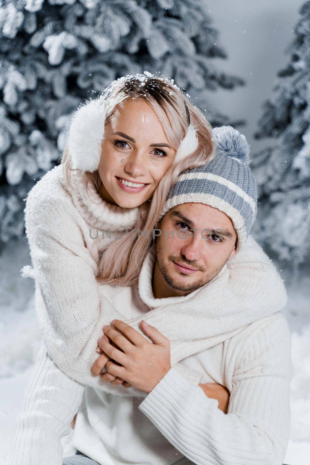 New year love story. Couple kisses and hugs. People weared wearing fur headphones, hats, white sweaters. Happy young couple hugs and kiss near christmas trees in winter day.