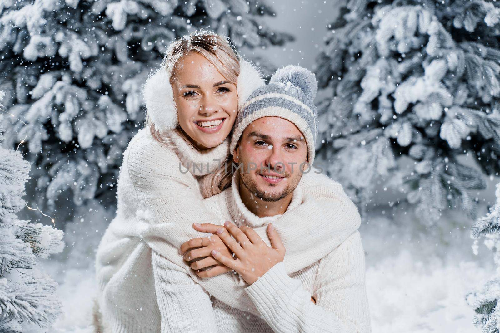 New year love story. Couple kisses and hugs. People weared wearing fur headphones, hats, white sweaters. Happy young couple hugs and kiss near christmas trees in winter day by Rabizo