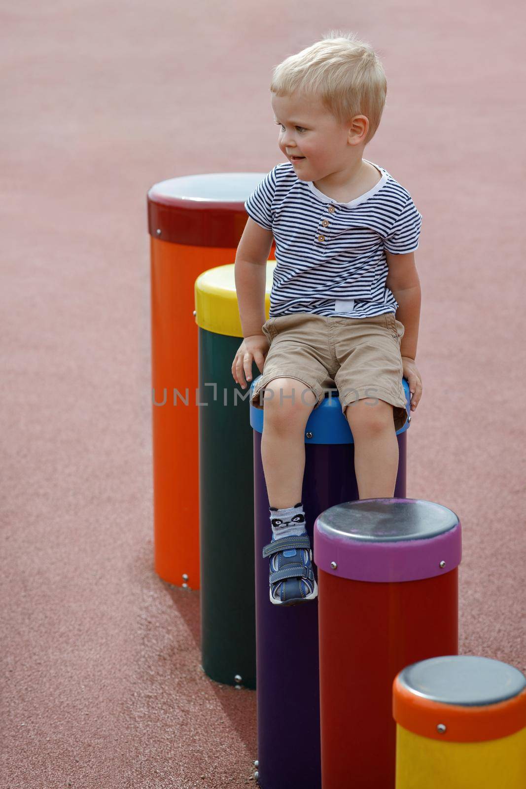 Cheeky kid is sitting on a line of bright colour drums in a modern public playground. Resting after an activities in summer time