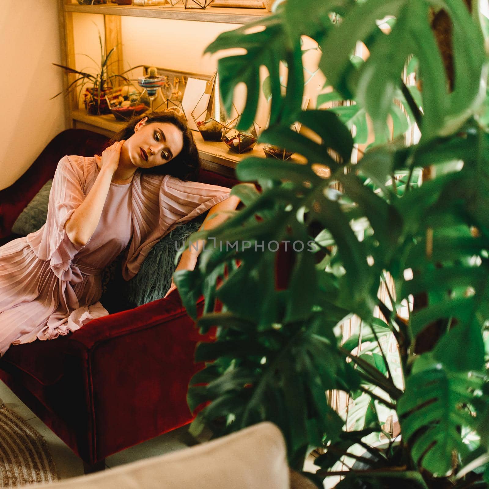 Dreamily girl is sitting ot the red sofa and think about a summer vacation. Summer lifestyle. Woman in the cafe with big green plants. by Rabizo